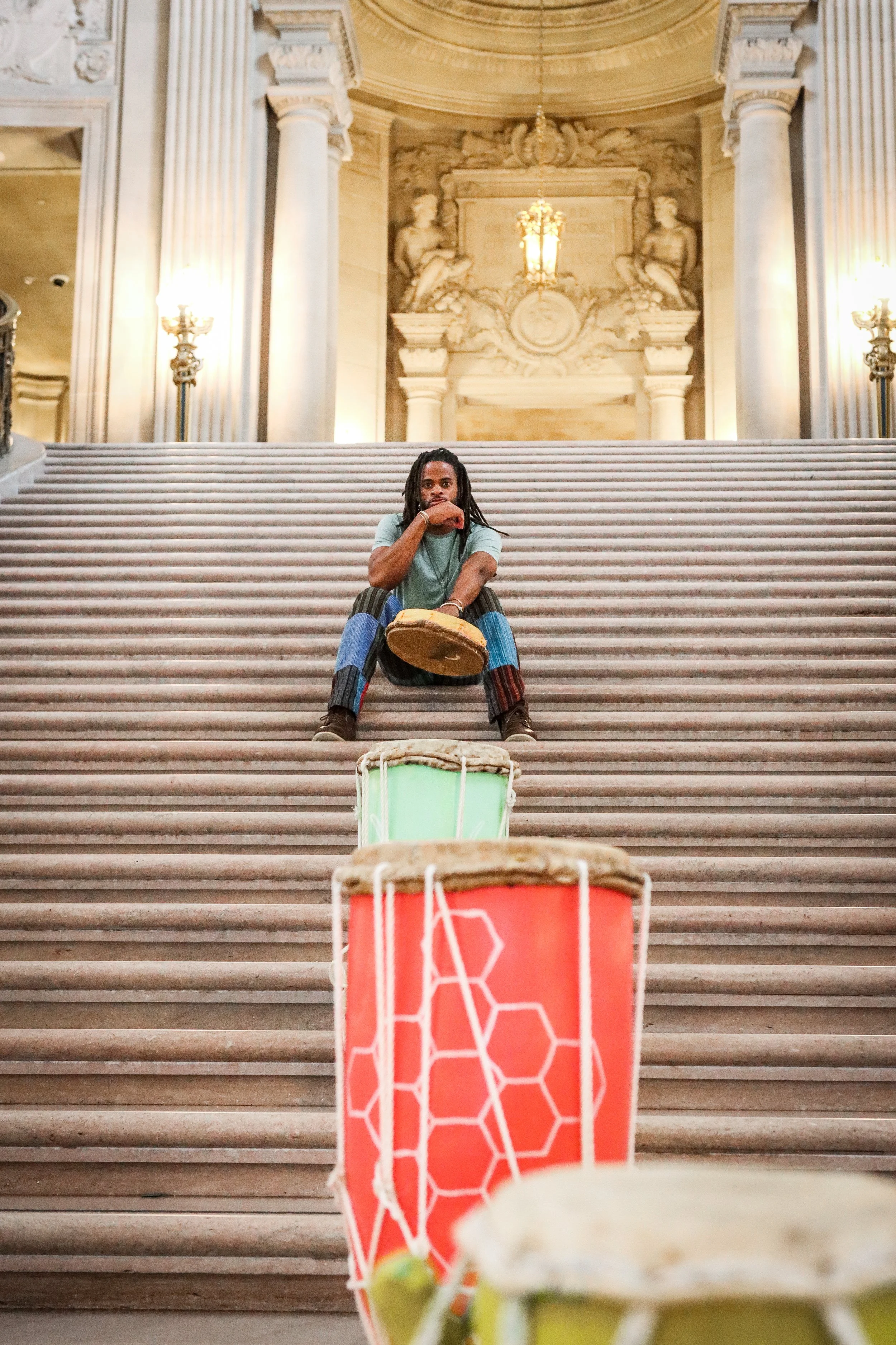 A person sitting on grand staircase of a historic building with ornate architecture, large columns, and statues, with drums and drums in the foreground.