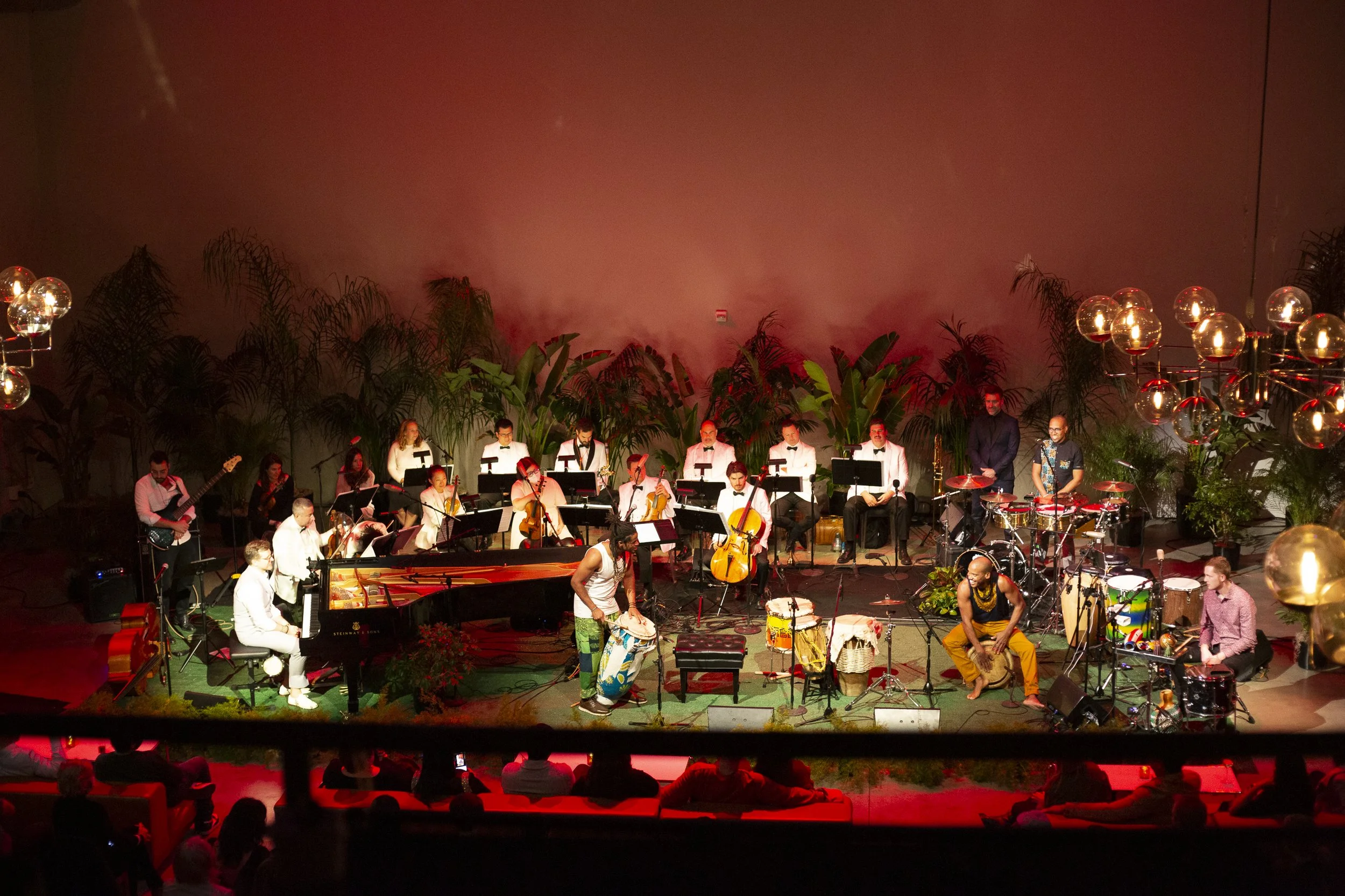 A large group of musicians on stage performing with various instruments, including violins, drums, a grand piano, and wind instruments, surrounded by lush green plants and decorative lighting, in an indoor concert setting.
