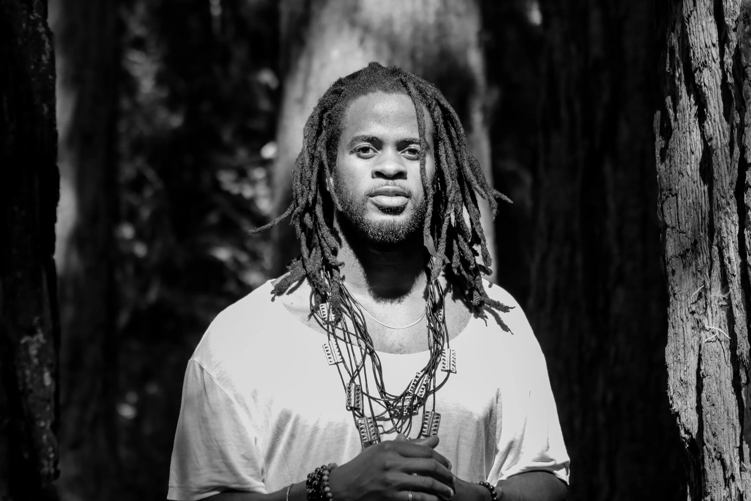 Black and white photo of a person with dreadlocks wearing a white shirt and layered necklaces, standing outdoors between two trees.