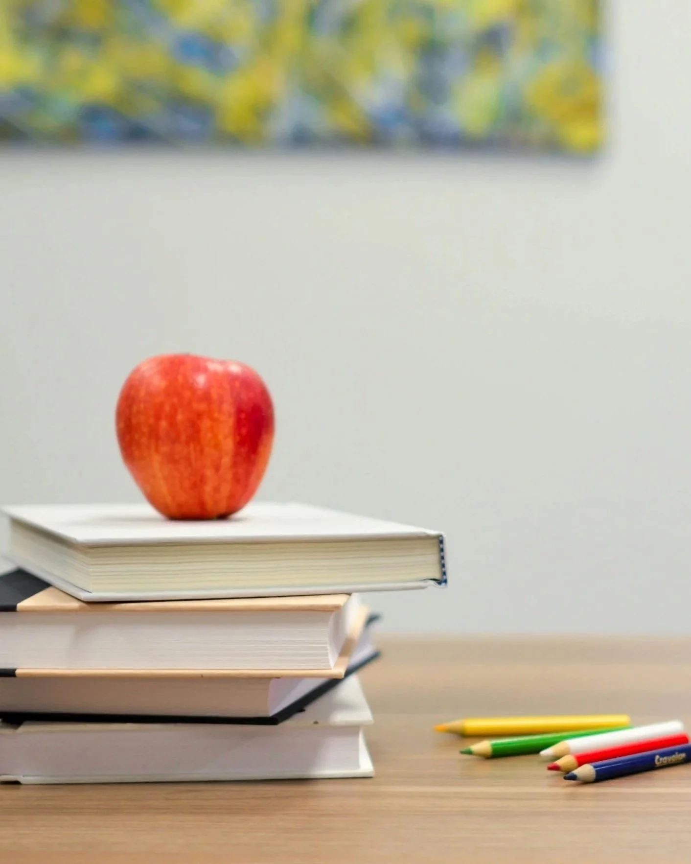 A red apple on top of a stack of three books on a wooden table, with colored pens on the right side of the table.