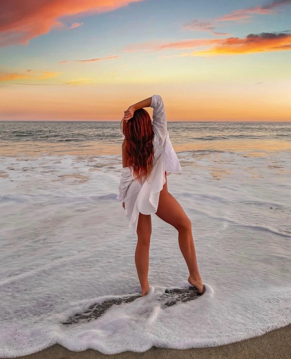 A woman with long red hair standing at the shoreline during a sunset, wearing a white shirt and no pants or shorts, with her hand on her head and the ocean waves around her feet.