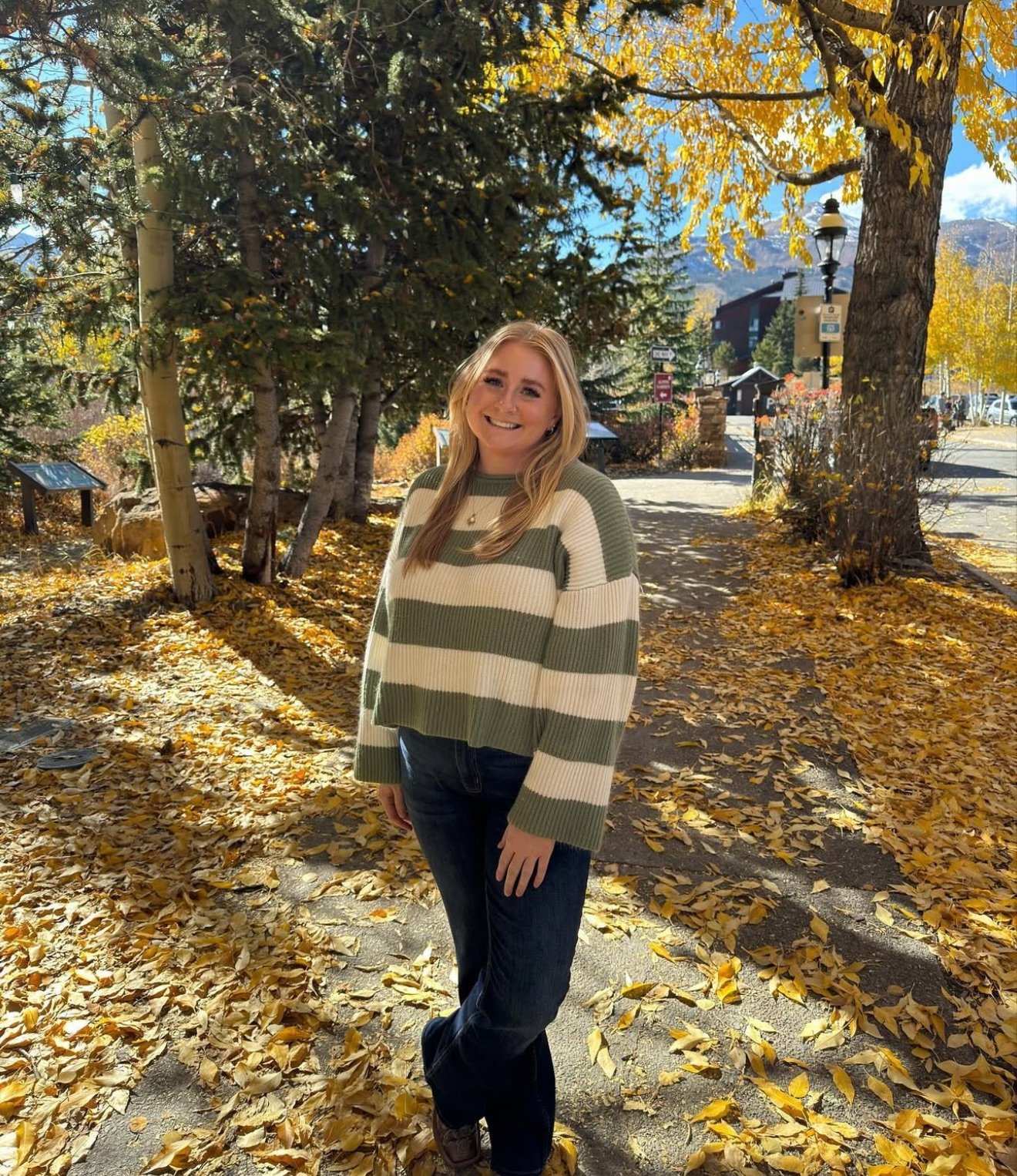 A woman with long blonde hair smiling wearing a green and white striped sweater and dark jeans standing on an autumn leaf-covered sidewalk park with trees and mountains in the background.