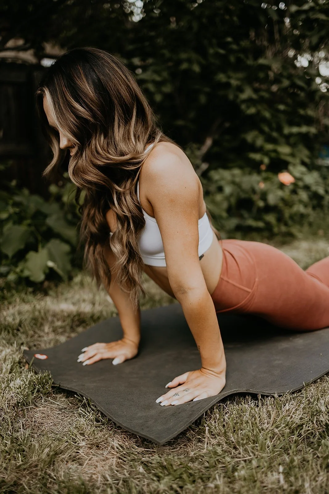 A woman with long, wavy brown hair performs a yoga pose on a black mat outdoors, surrounded by greenery.
