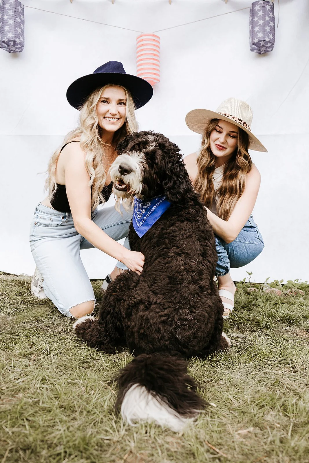 Two women with long hair, wearing hats and casual clothing, are squatting on the grass with a large, curly-haired dog that has a blue bandana around its neck. They are smiling and posing for the photo with a decorative background of hanging paper lan