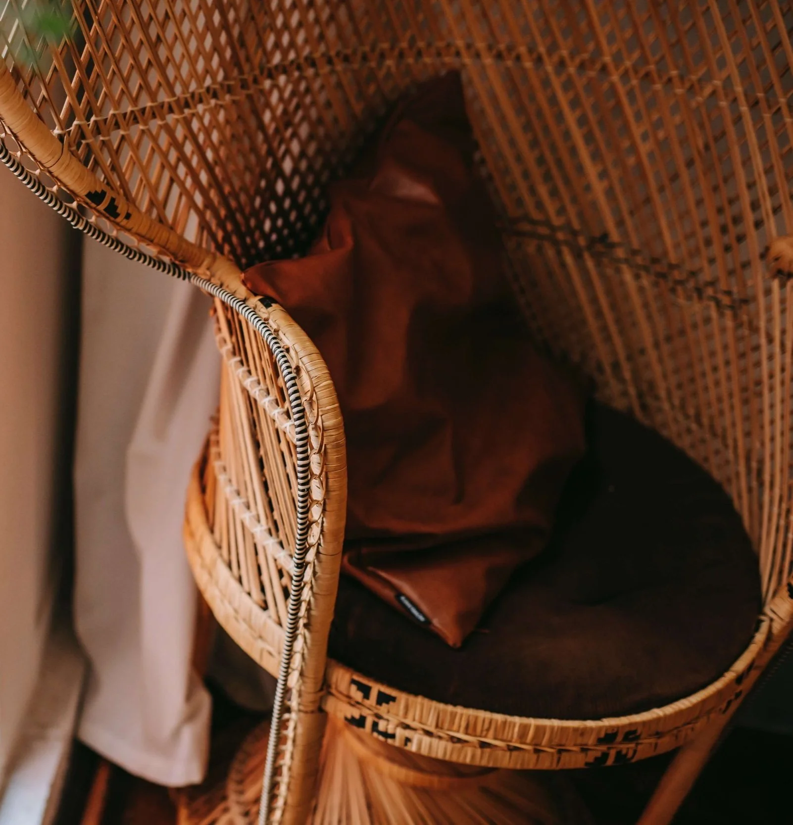 A wicker hanging chair with a brown cushion inside, placed against a wall with a curtain nearby.