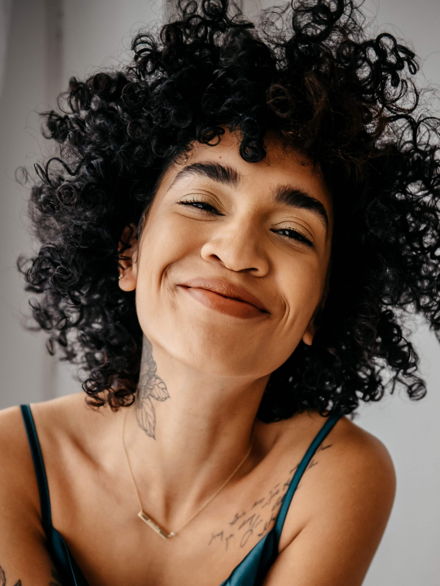 A woman with curly black hair smiling and wearing a teal spaghetti strap top, a gold necklace, and a tattoo of leaves on her neck and shoulder.