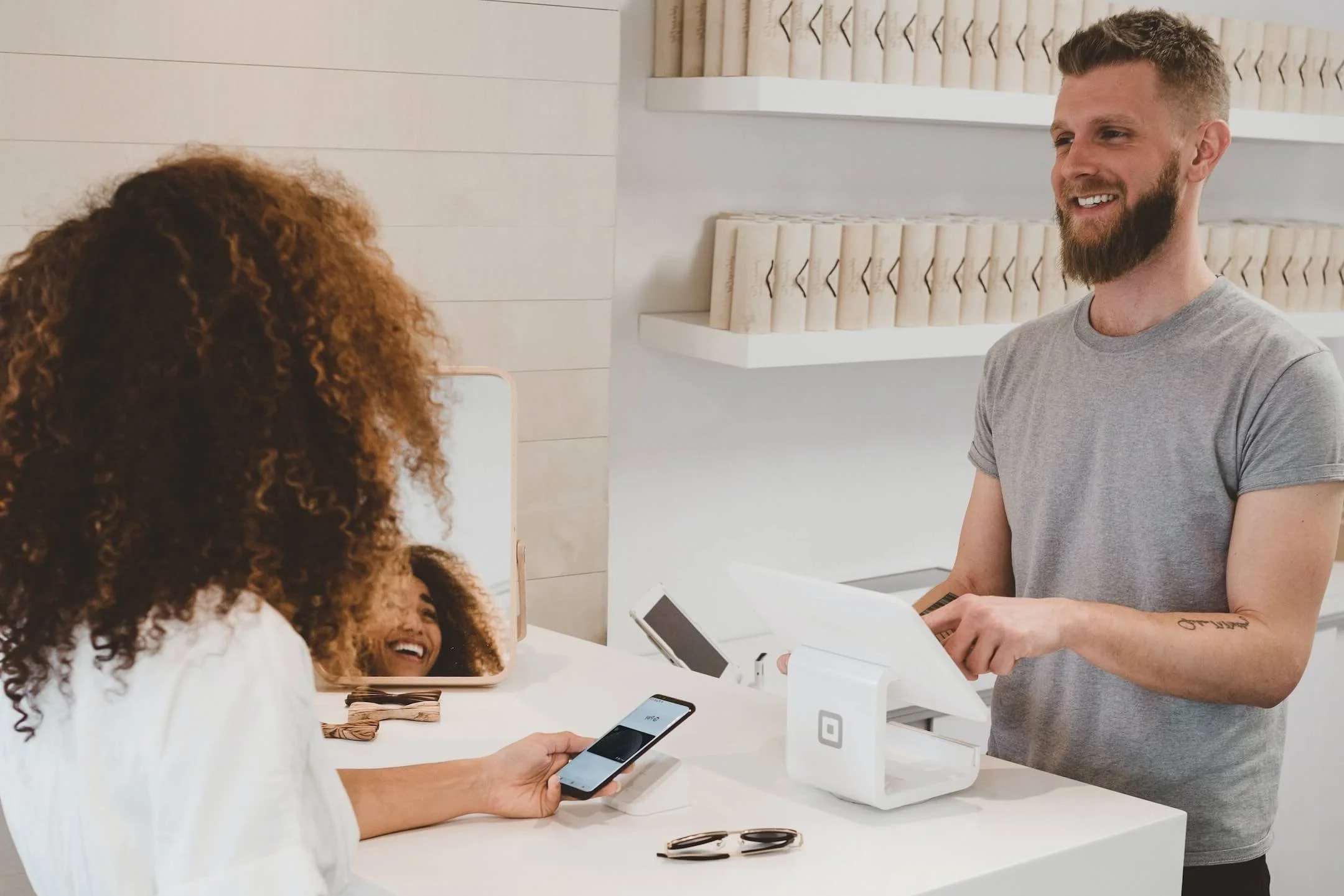 A person with a beard and short hair wearing a gray t-shirt is standing at a counter, smiling while using a payment terminal. A woman with curly hair, holding a smartphone, is smiling and looking at the man. Another woman with long curly hair is visi