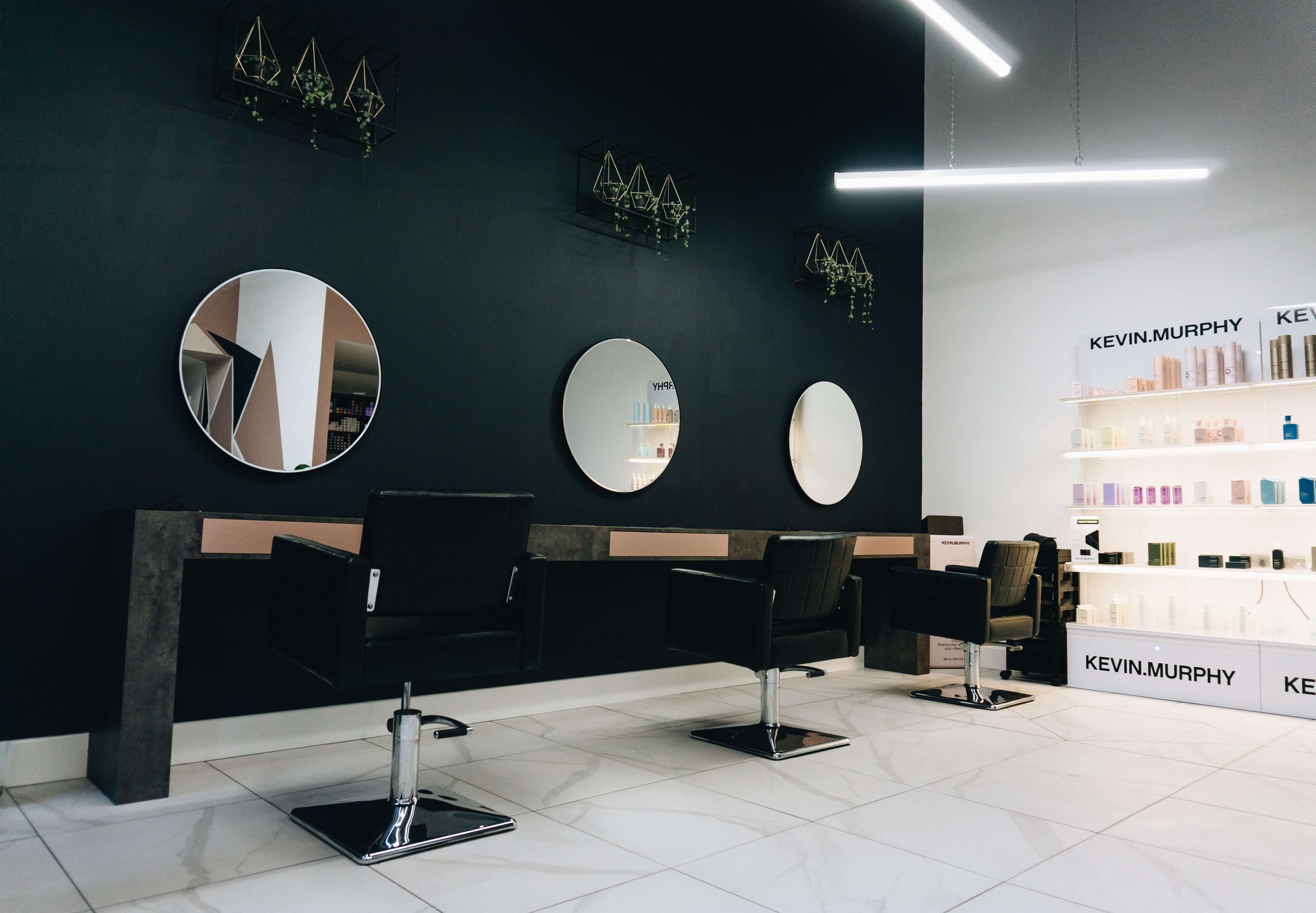 Salon interior with black chairs, three circular mirrors on dark wall, white shelving with beauty products, and large bright fluorescent lights.