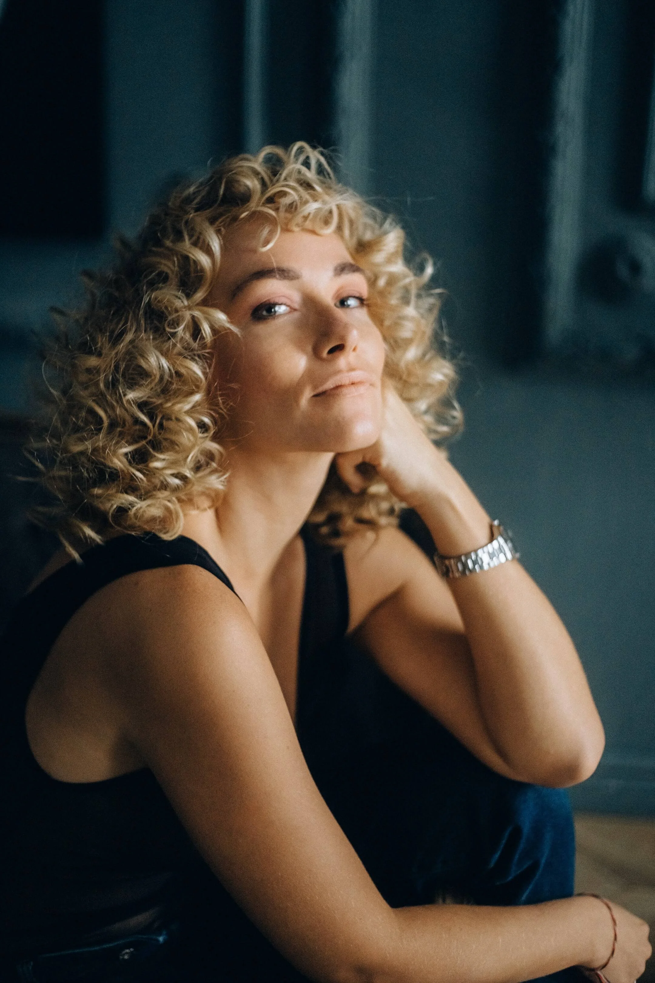 A woman with curly blonde hair is sitting and looking slightly upwards, resting her chin on her hand, with a wristwatch on her wrist, against a dark background.