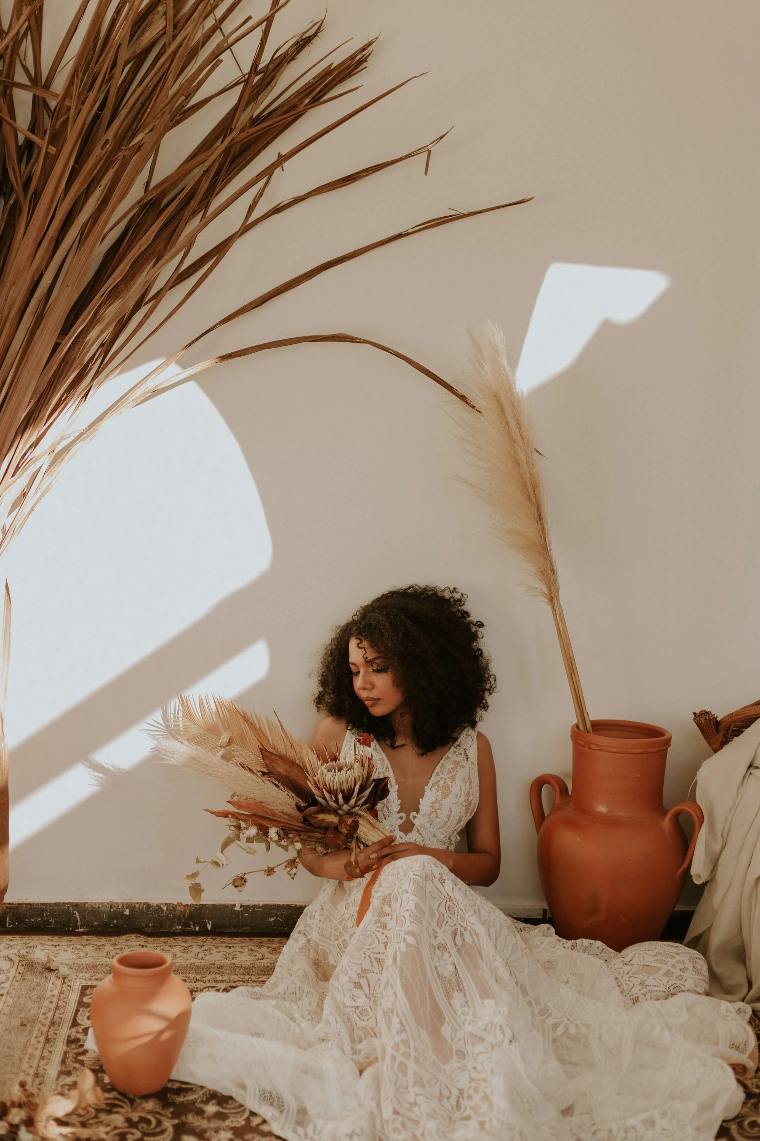 A woman with curly hair sitting on the floor in front of a white wall, holding a bouquet of dried flowers and plants, with large terracotta vases and a patterned rug around her.