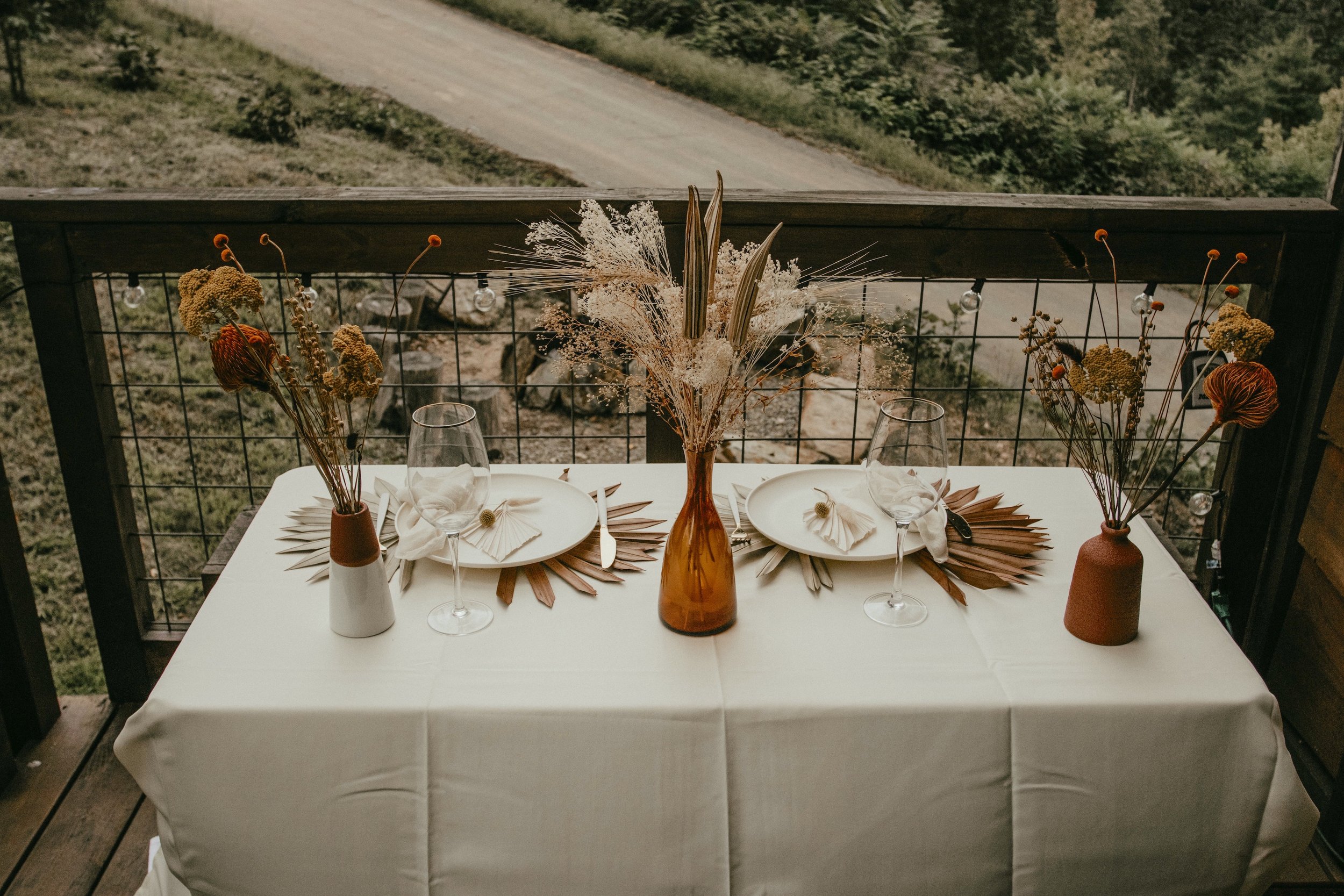 Table set for outdoor dining with a white tablecloth, decorative paper fans, and glassware, featuring a central bouquet of dried flowers in amber and white vases, on a wooden balcony overlooking a scenic rural landscape.