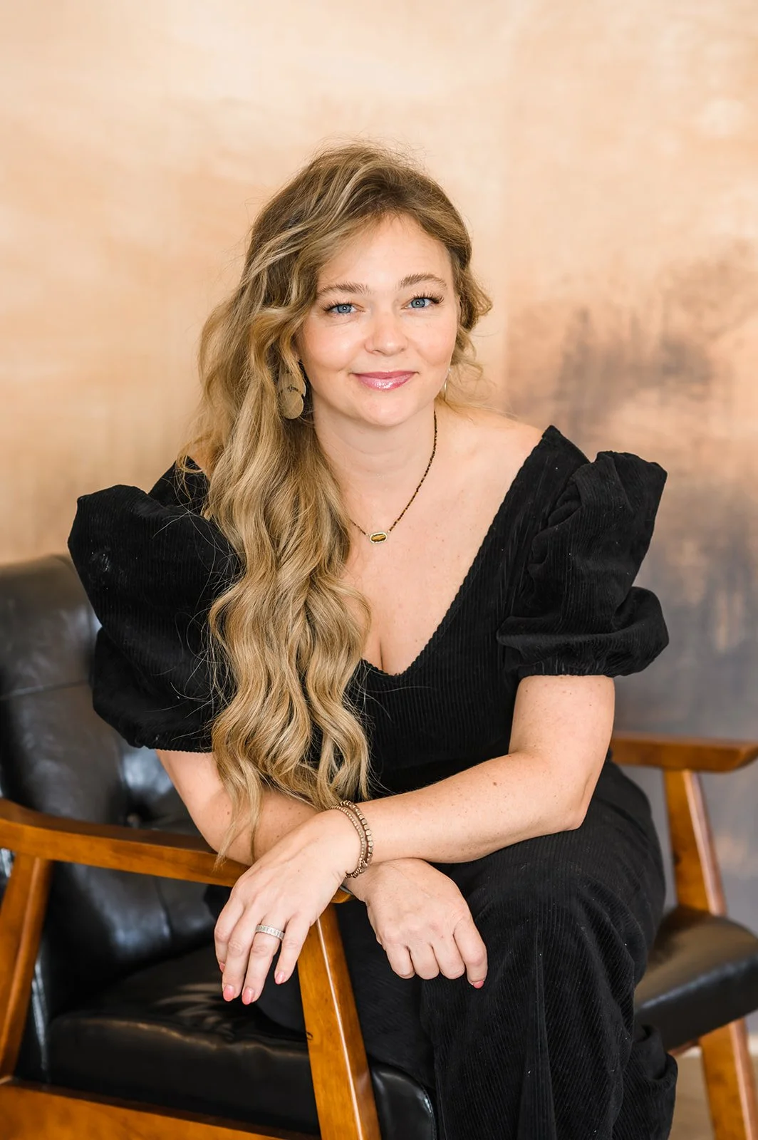 A woman with long wavy blonde hair and blue eyes, wearing a black dress with puffed sleeves, seated on a wooden chair with black leather cushioning, posed against a warm, textured beige wall.