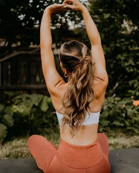 Woman practicing yoga outdoors in a garden with her arms raised overhead and back facing the camera.