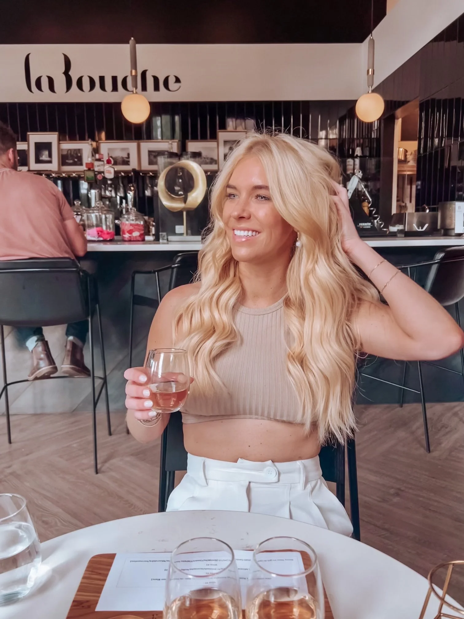 A blonde woman with long wavy hair smiling, holding a glass of water in a restaurant or cafe. She is wearing a beige crop top and white pants. In the background, there is a bar with various bottles and glasses, and a man wearing a pink shirt sitting 