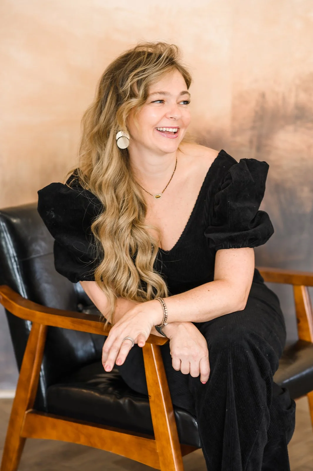 A woman with long, wavy blonde hair wearing a black dress with puffed sleeves, gold jewelry, and sitting on a wooden chair with a black leather seat, smiling and looking to her right.