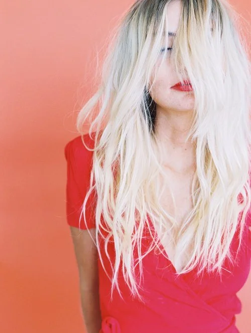 Woman with long, wavy blonde hair wearing a red top against a pink background.