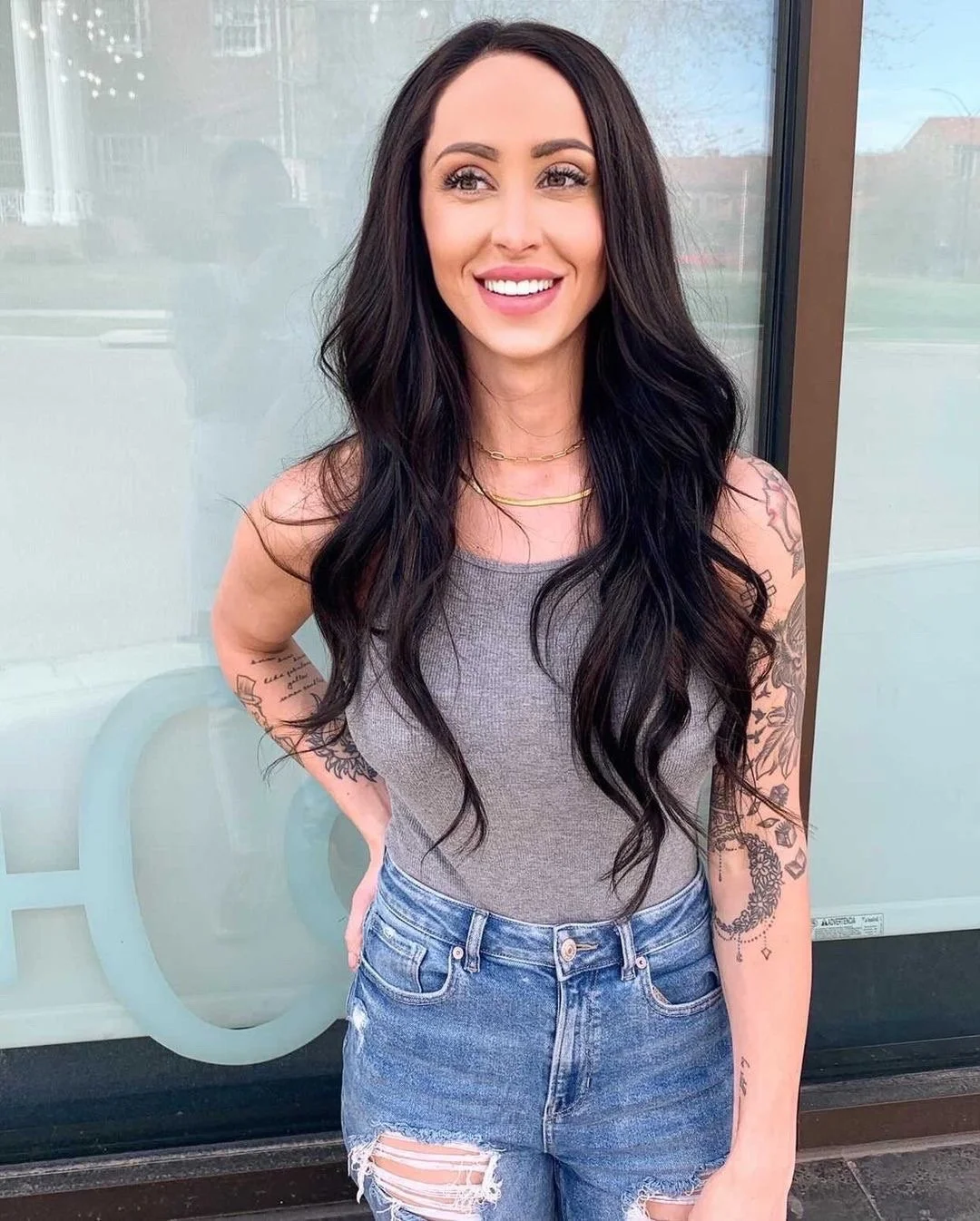 A woman with long dark wavy hair, wearing a gray sleeveless top and ripped denim shorts, smiling outdoors.
