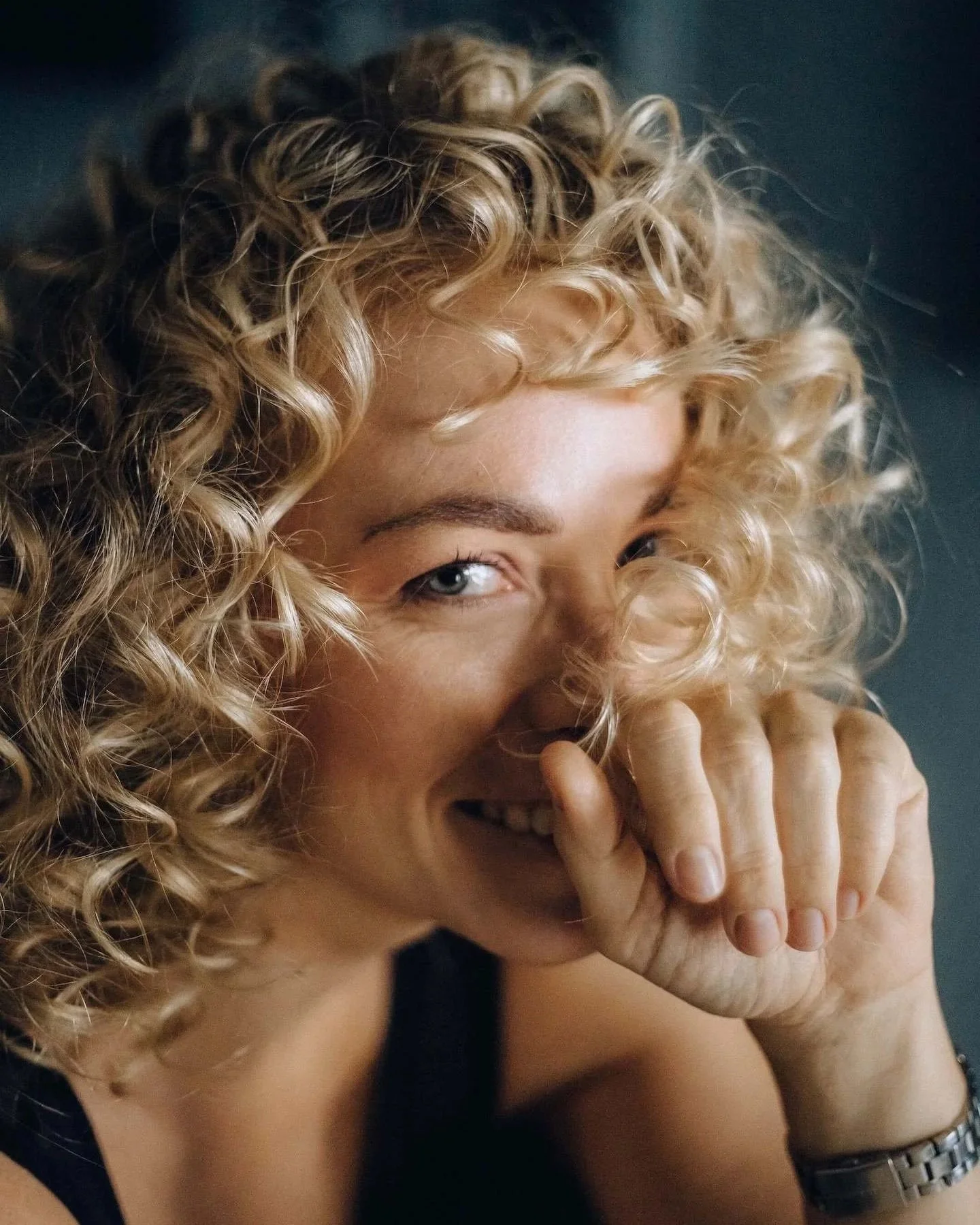 A woman with curly blonde hair smiling and covering her mouth with her hand.
