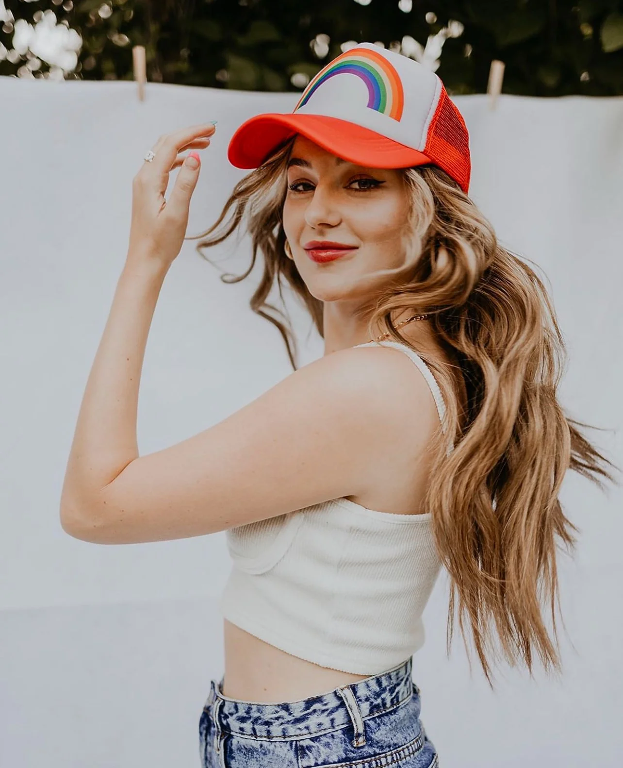 Young woman with long wavy hair wearing a red and white trucker hat with a rainbow, white sleeveless crop top, and high-waisted patterned jeans, standing against a white background.