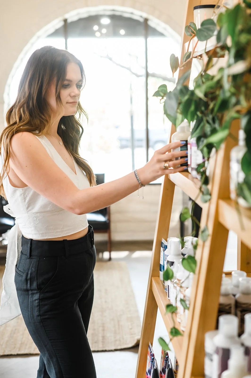 A woman with long brown hair wearing a white sleeveless top and black pants shopping for skincare products in a store with white shelves and green plants.