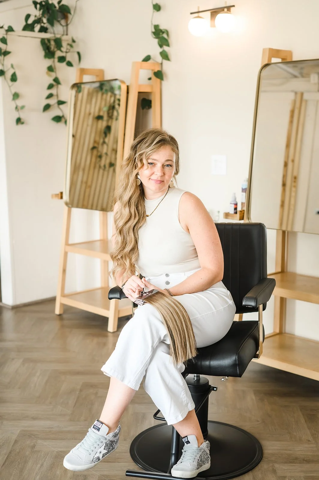 A woman with long wavy blonde hair sitting on a black salon chair, holding a hair sample, in a hair salon with a mirror, plant, and wooden ladder in the background.