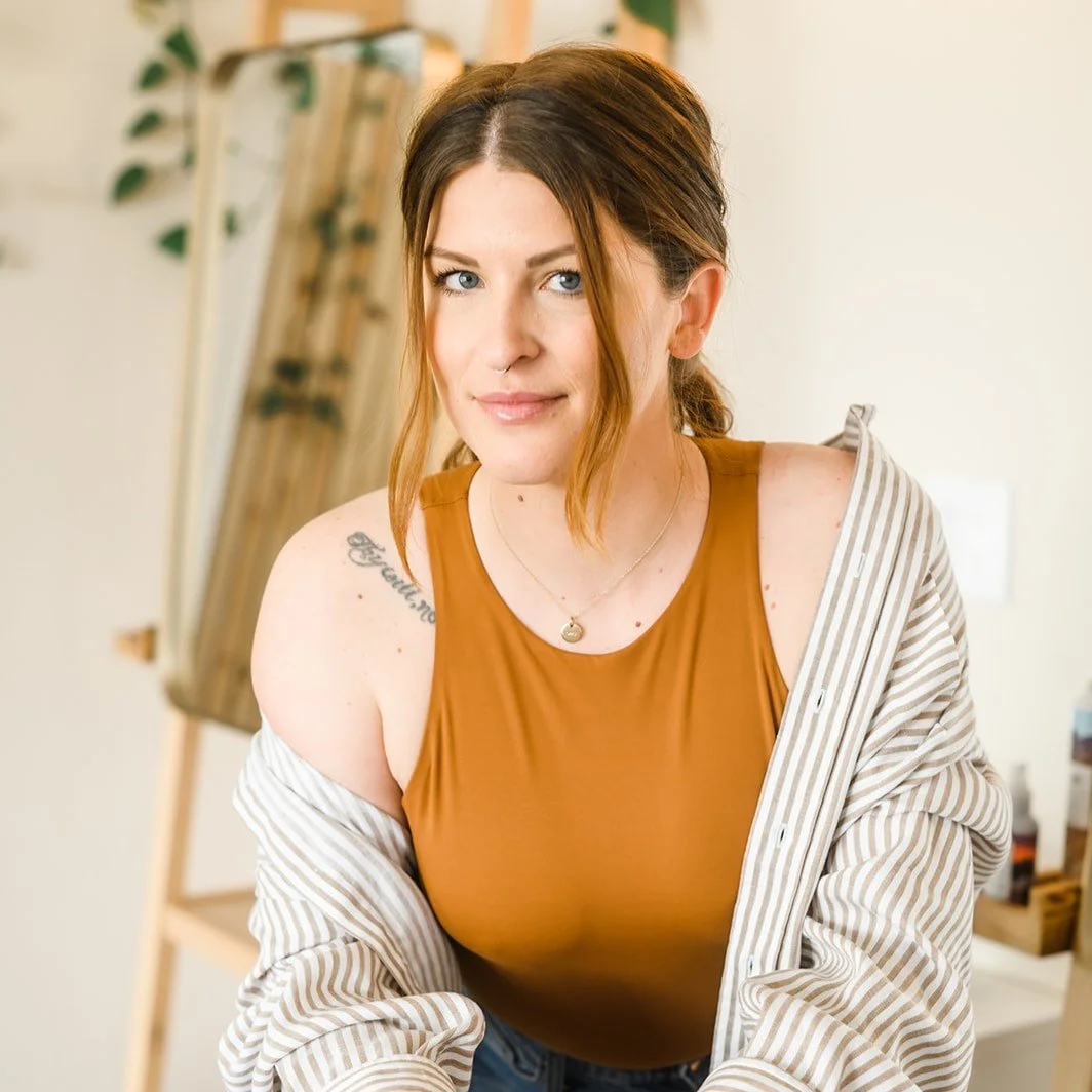 A woman with brown hair and blue eyes wearing a brown tank top and striped shirt, standing indoors with a mirror and plant in the background.