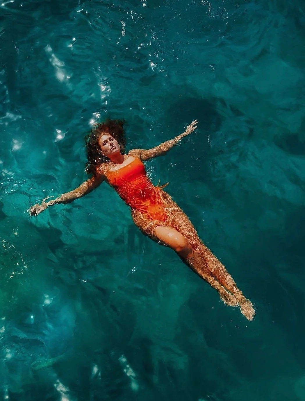 A woman floating on her back in a swimming pool, wearing an orange swimsuit and beige sheer cover-up, with her arms outstretched and hair spread out in the water.