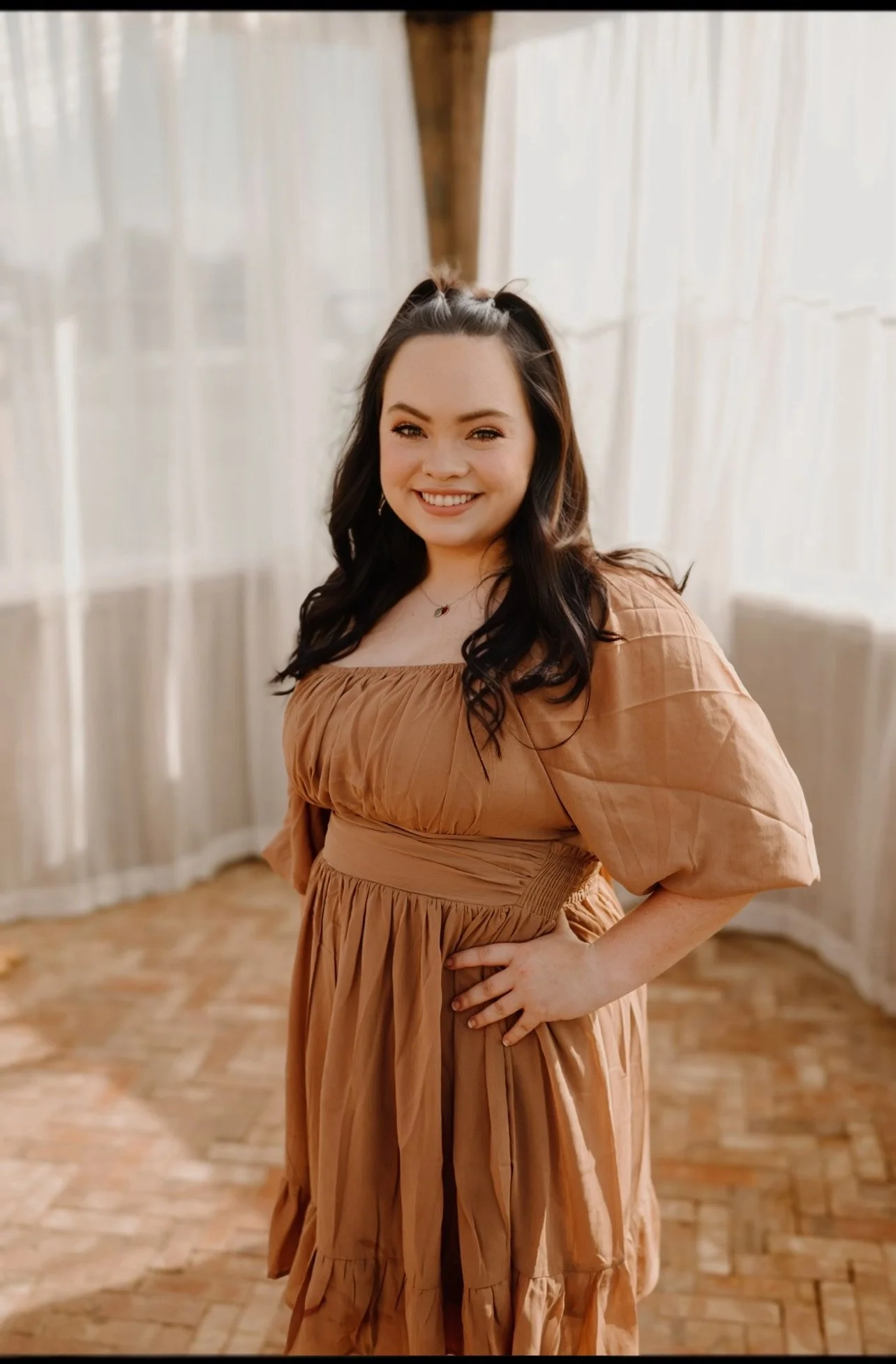 Young woman with dark hair and a smile, wearing a tan dress with puffy sleeves, standing in a well-lit room with white curtains and a patterned wooden floor.