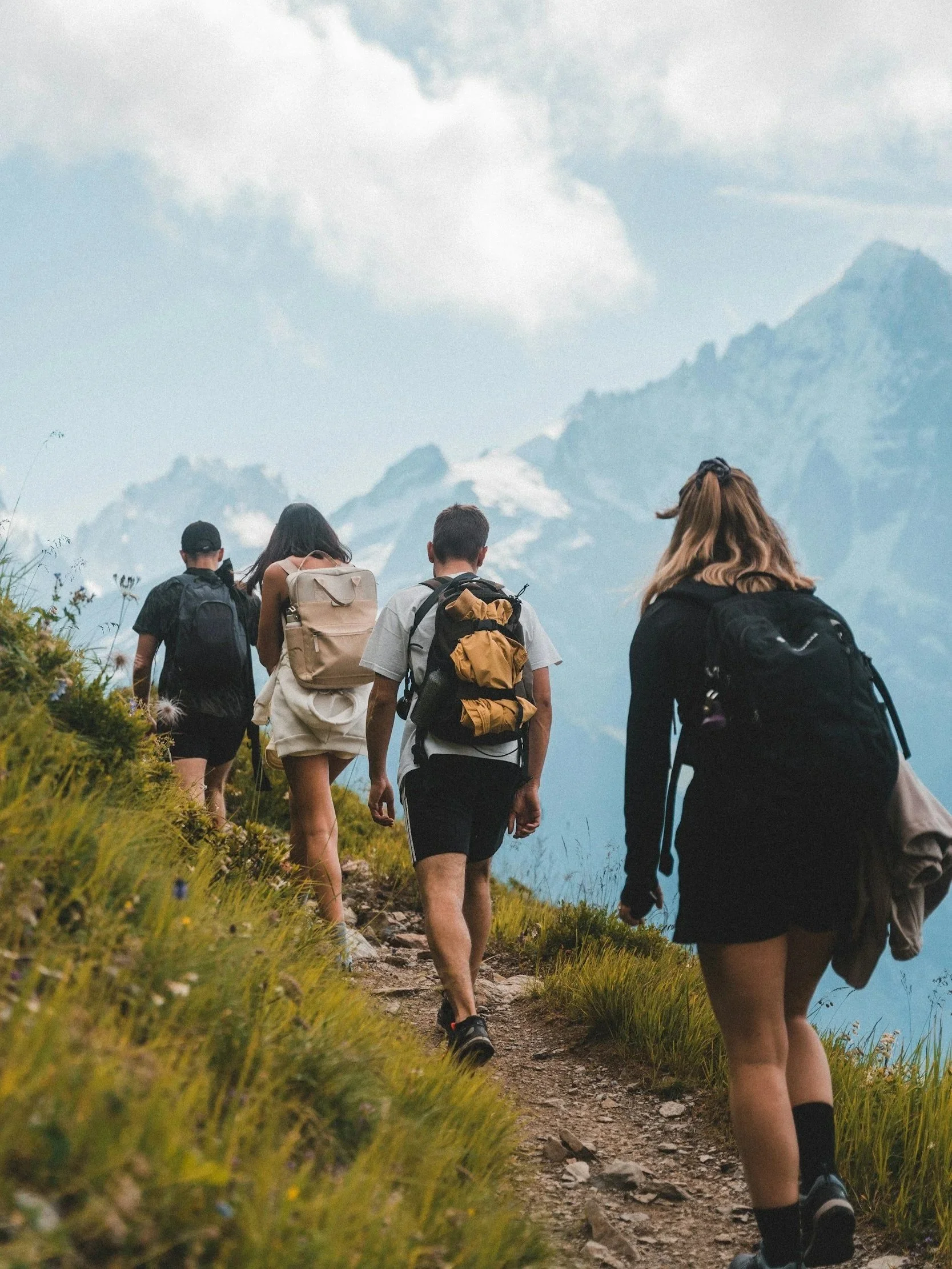 Four hikers walking on a rocky mountain trail with a mountain range and cloudy sky in the background.