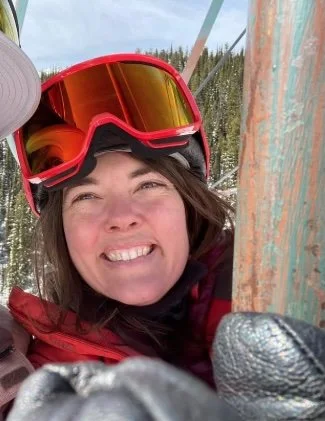 A woman wearing ski goggles and a red jacket smiling outdoors near a ski lift.