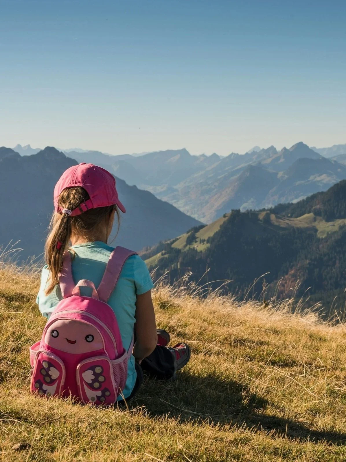 Young girl with a pink cap and backpack sitting on grassy hillside overlooking mountainous landscape.