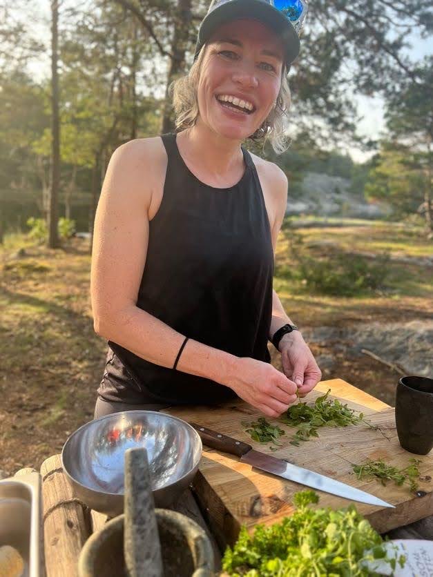 A woman with a black tank top and a cap smiles while preparing herbs outdoors at a wooden table.