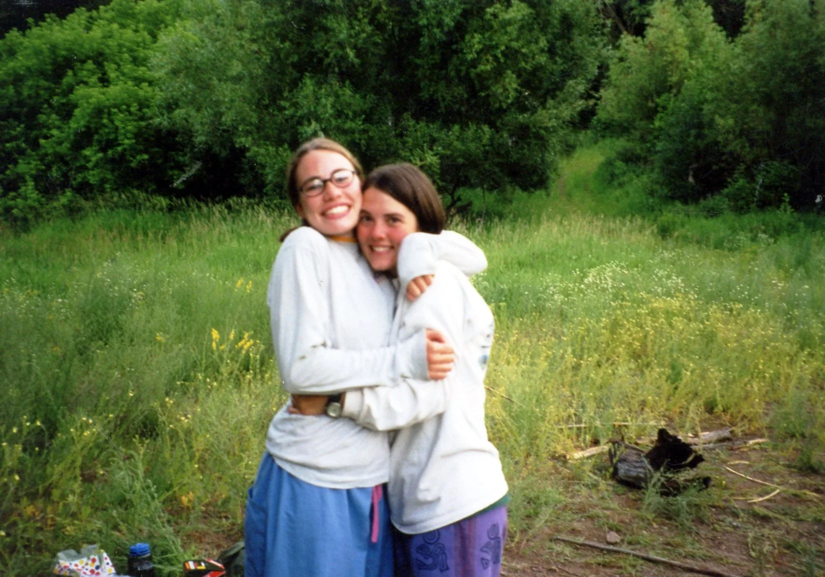 Two women hugging and smiling outdoors in a green, grassy field with trees in the background.