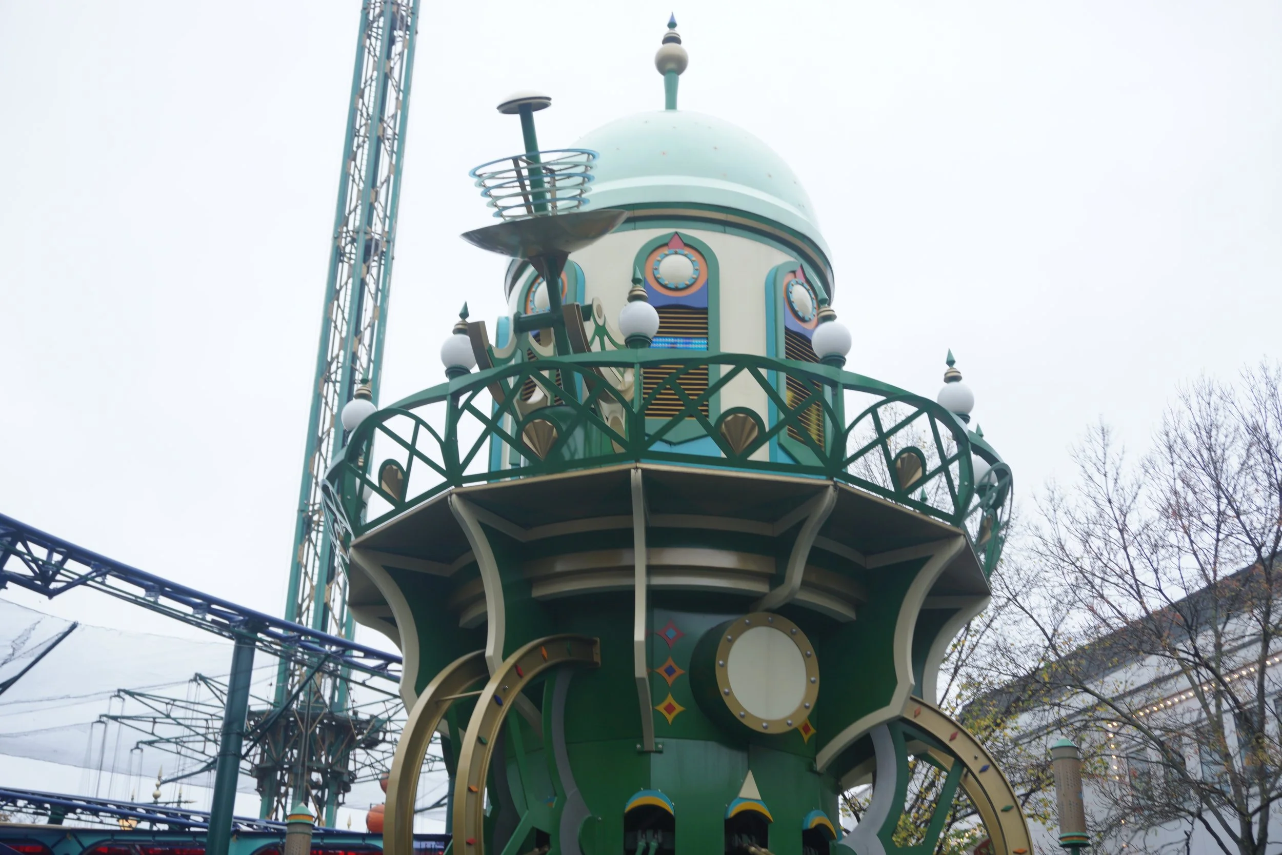 A whimsical, colorful amusement park ride station with a spaceship or rocket theme, featuring a green and cream color scheme and decorative accents, set against a cloudy sky.