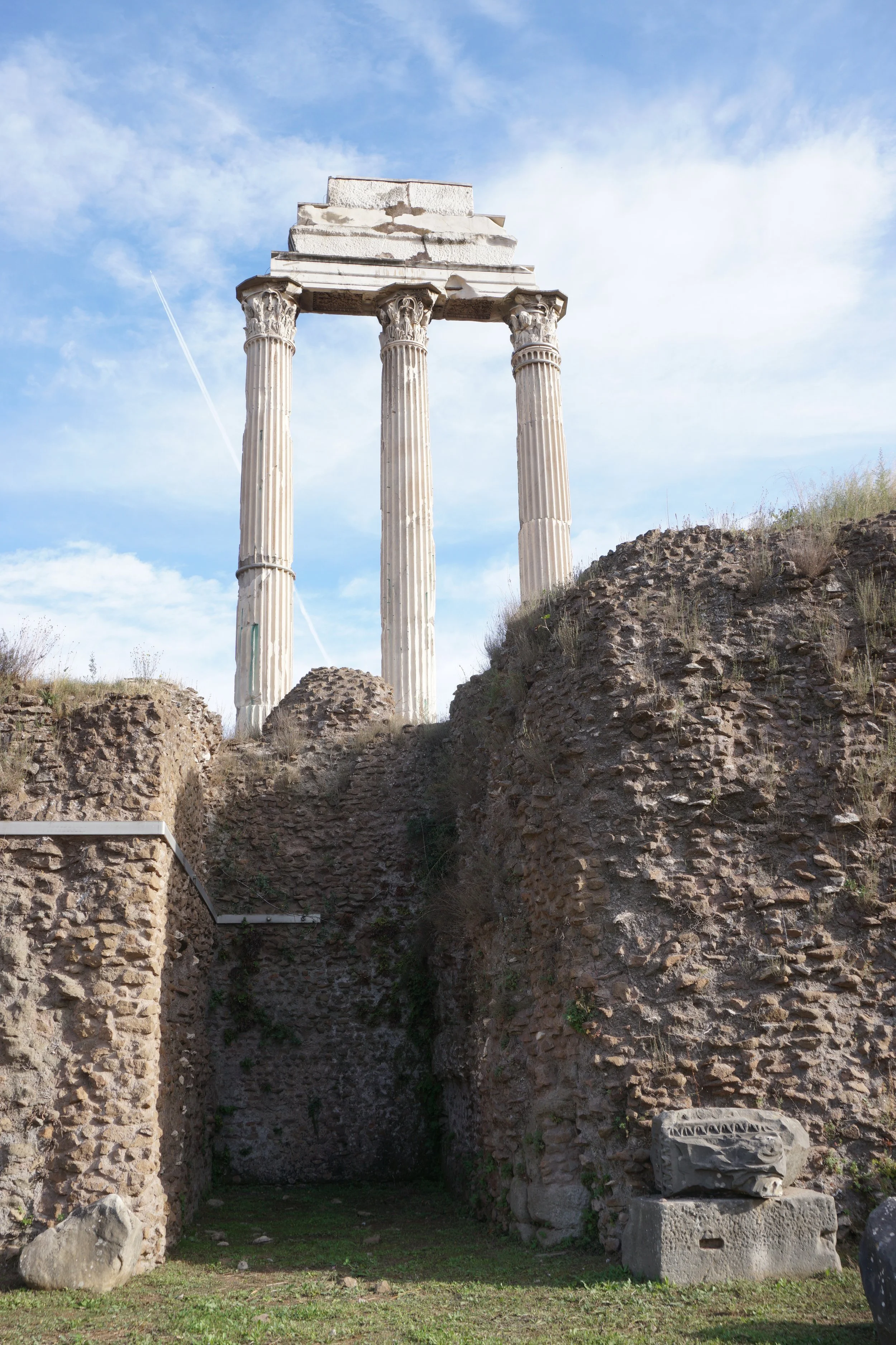 Ancient Roman ruins with three standing columns and a broken arch, set against a blue sky with wispy clouds.