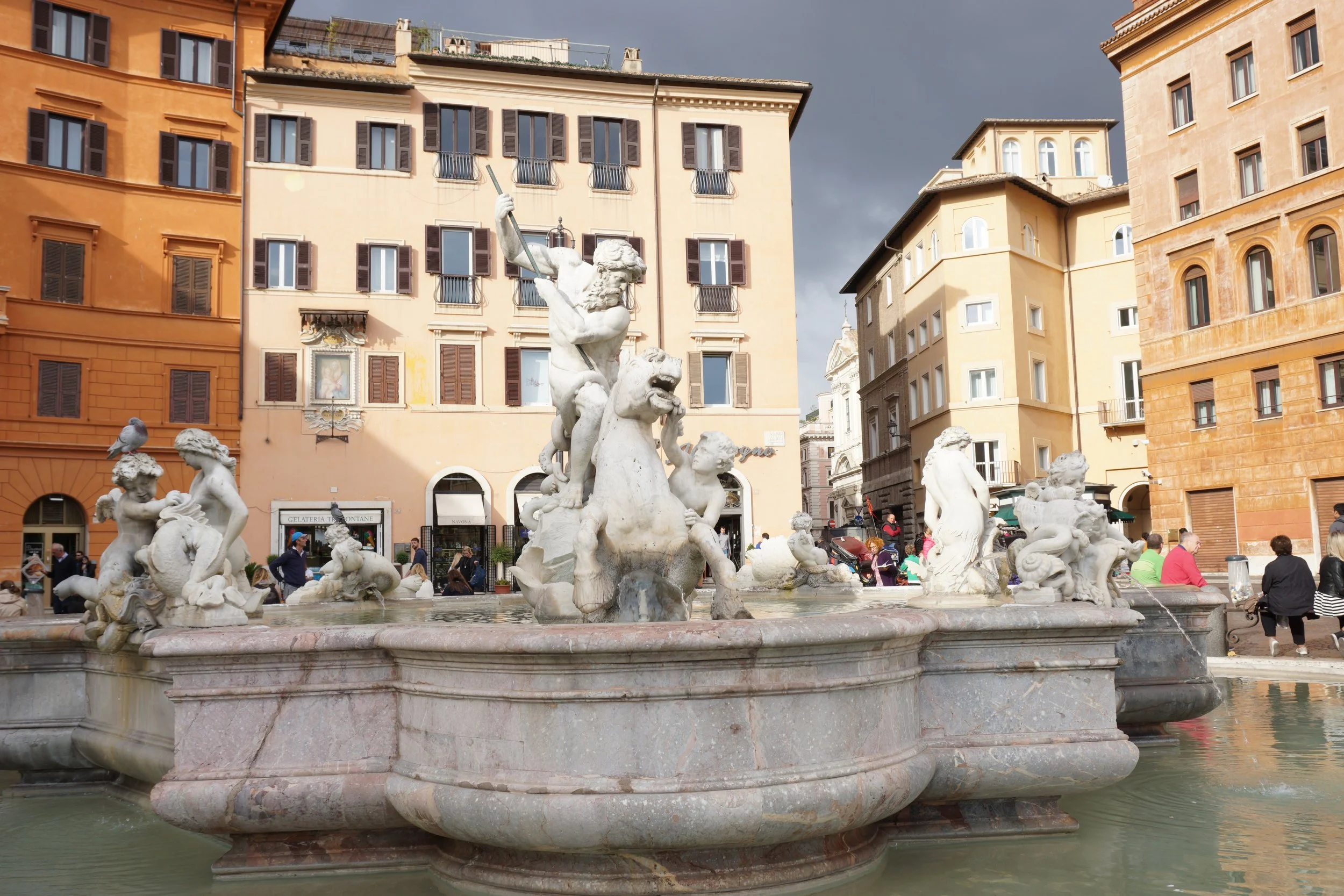 A fountain with multiple marble sculptures of mythological figures in a city square, surrounded by colorful buildings and people walking and sitting around.