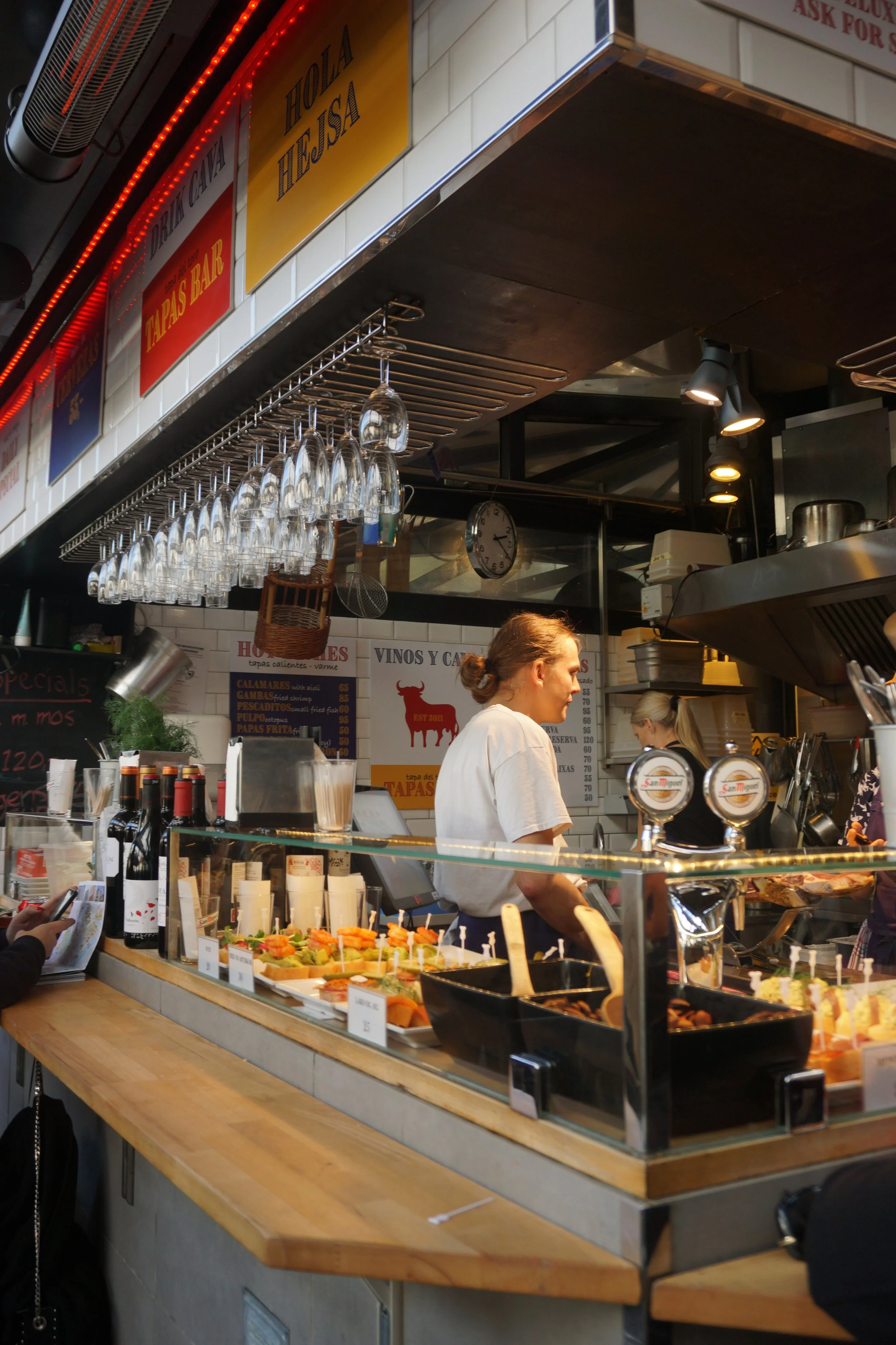 Inside of a tapas bar with hanging wine glasses, a menu, and a woman working behind the counter preparing food.