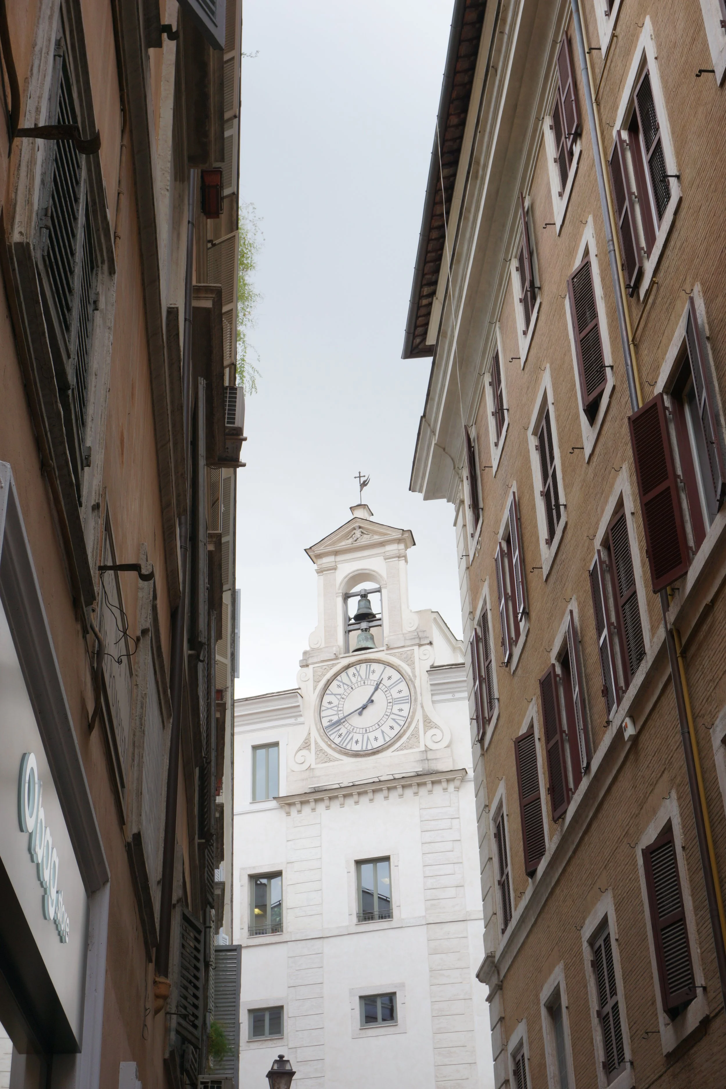 View of a clock tower with a cross on top, seen through a narrow street flanked by tall multi-story buildings with windows and shutters.
