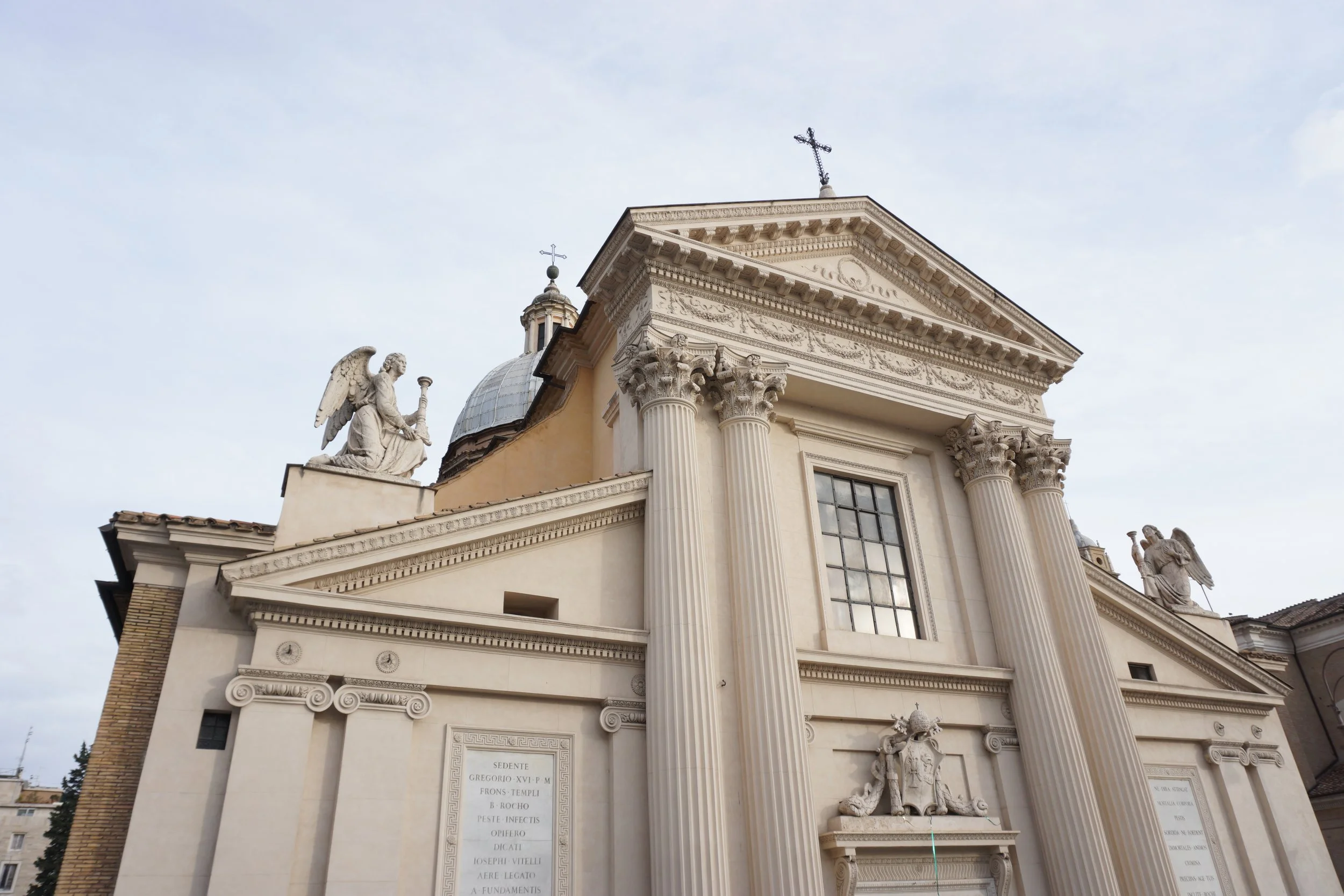 Front view of a historic church with classical columns, sculptures of angels on the roof, and a cross on the top.