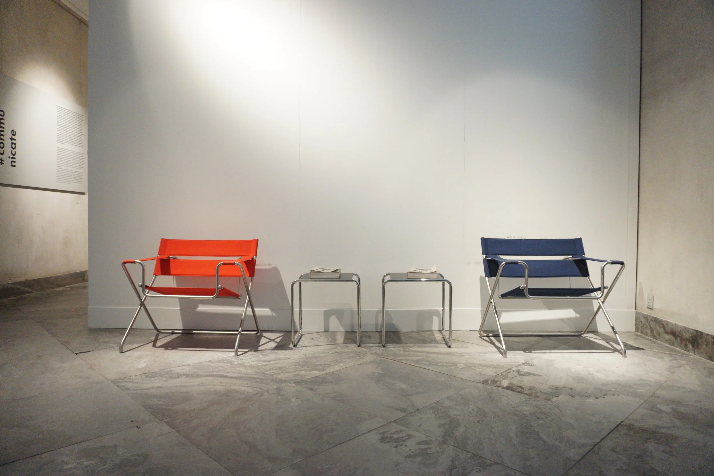 Two modern chairs, one orange and one blue, are placed on either side of two small tables with newspapers on top, against a white wall in a contemporary interior setting.