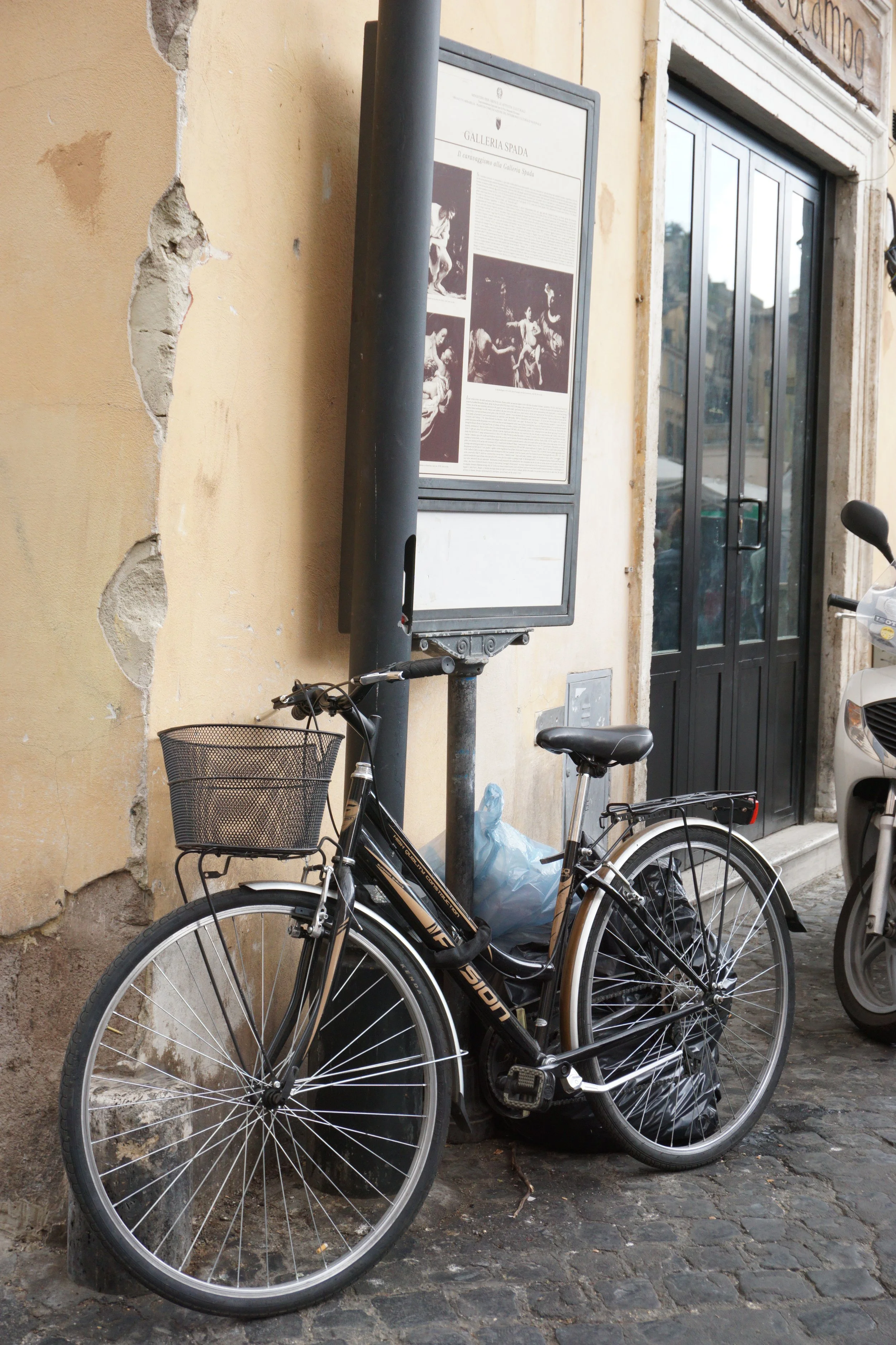 A bicycle with a basket parked against a yellow, distressed wall near a black pole and display board outside a building entrance.