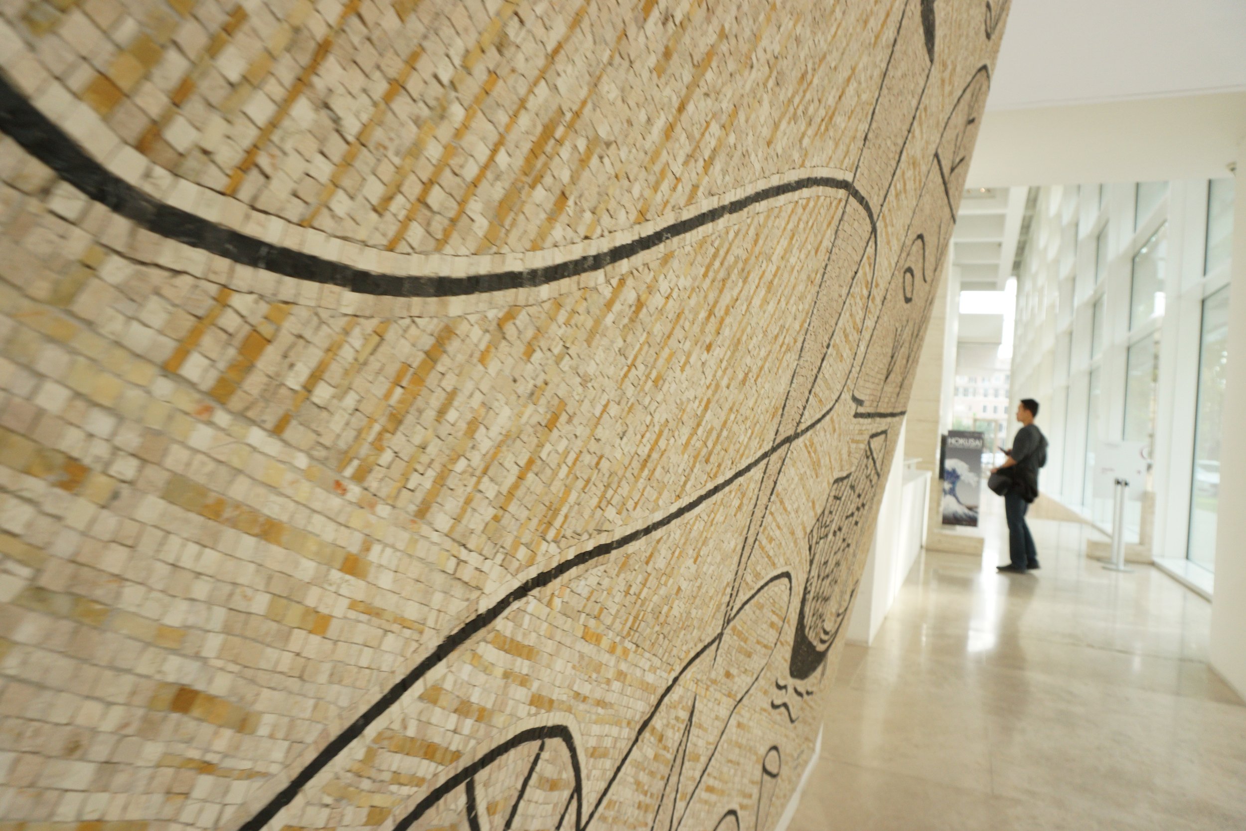A person standing in front of a large wall mosaic depicting a map or diagram, inside a modern building with large windows.