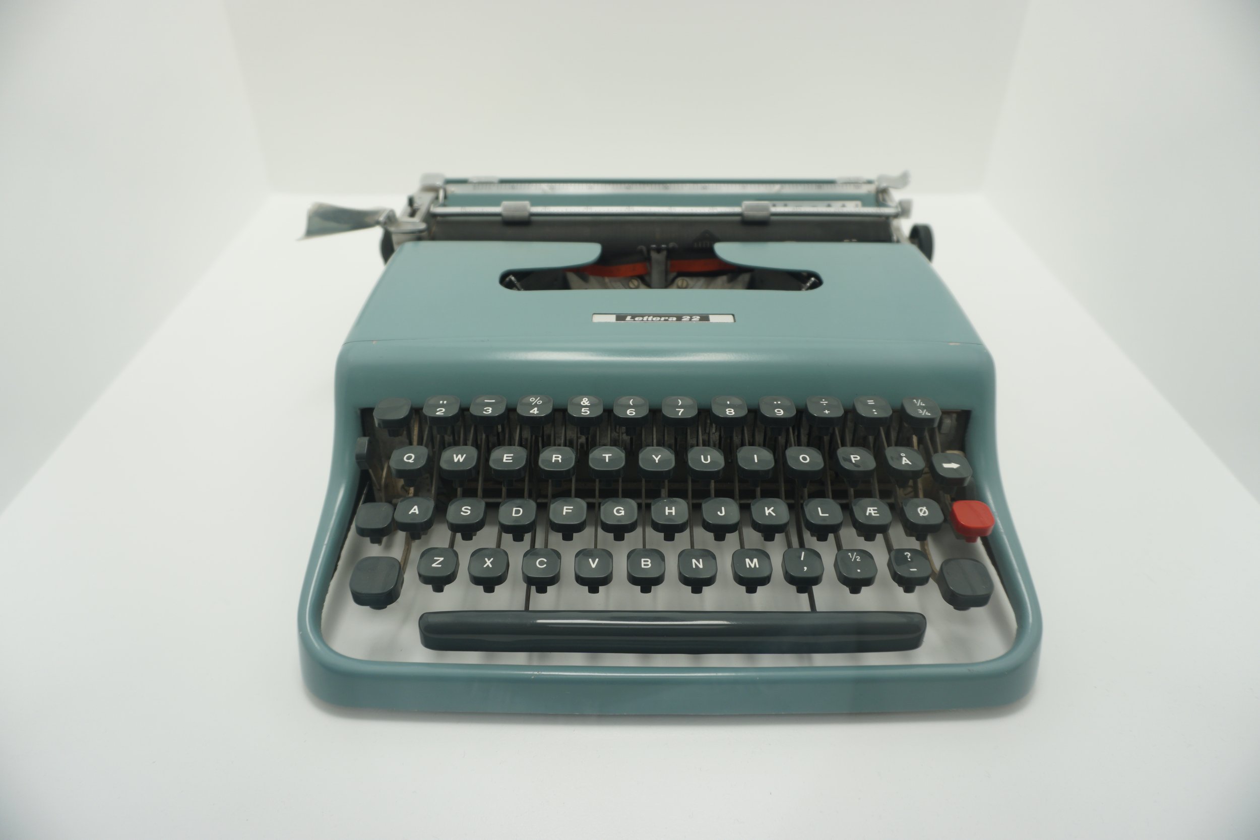 A vintage turquoise typewriter with black keys and a red key on the right side, placed on a white surface.