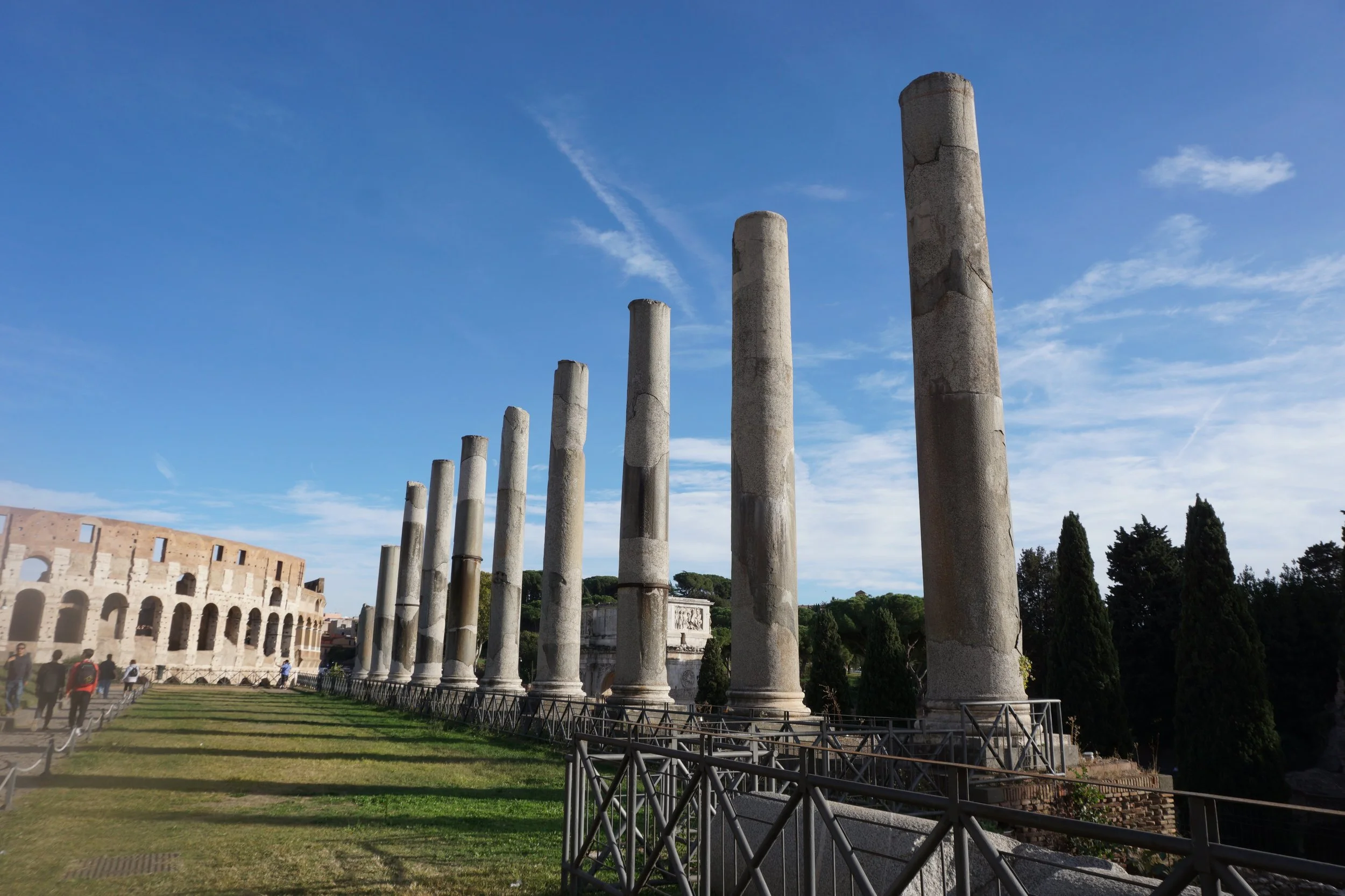 Ancient Roman ruins with nine tall weathered stone columns lined up in a row, with the Colosseum visible in the background under a bright blue sky.