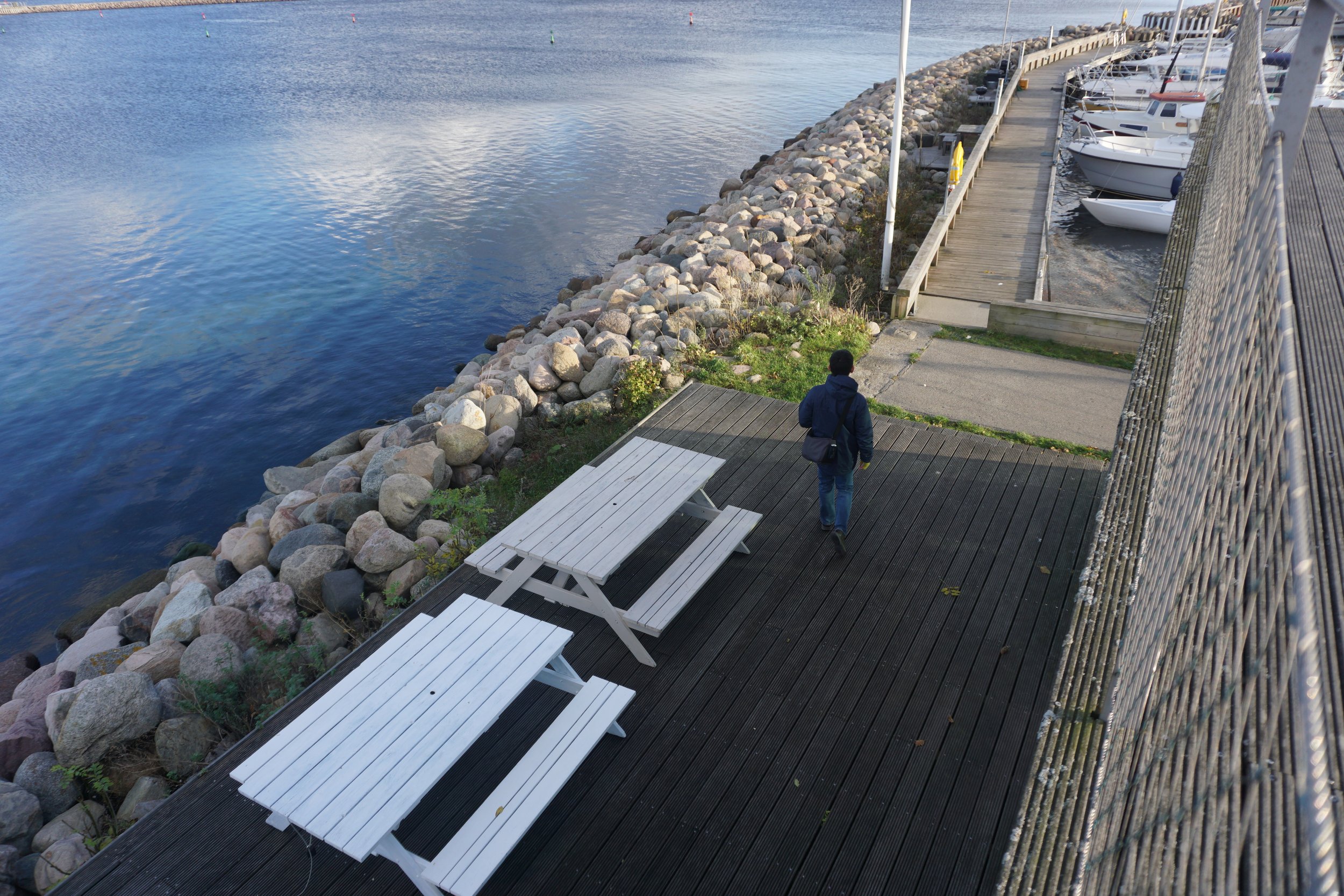 A person walking along a wooden dock beside a marina, with boats docked on the right and a lake on the left. There are two white benches on the dock, and a rocky shoreline separating the water from the land.