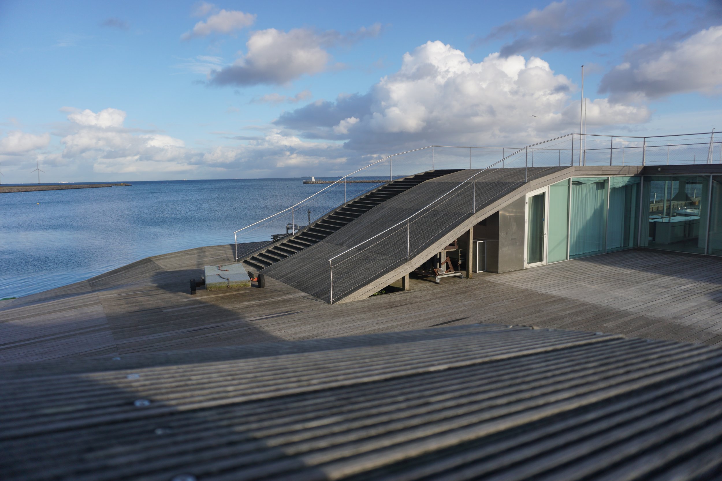 Modern building with a rooftop staircase, overlooking a body of water under a partly cloudy sky.