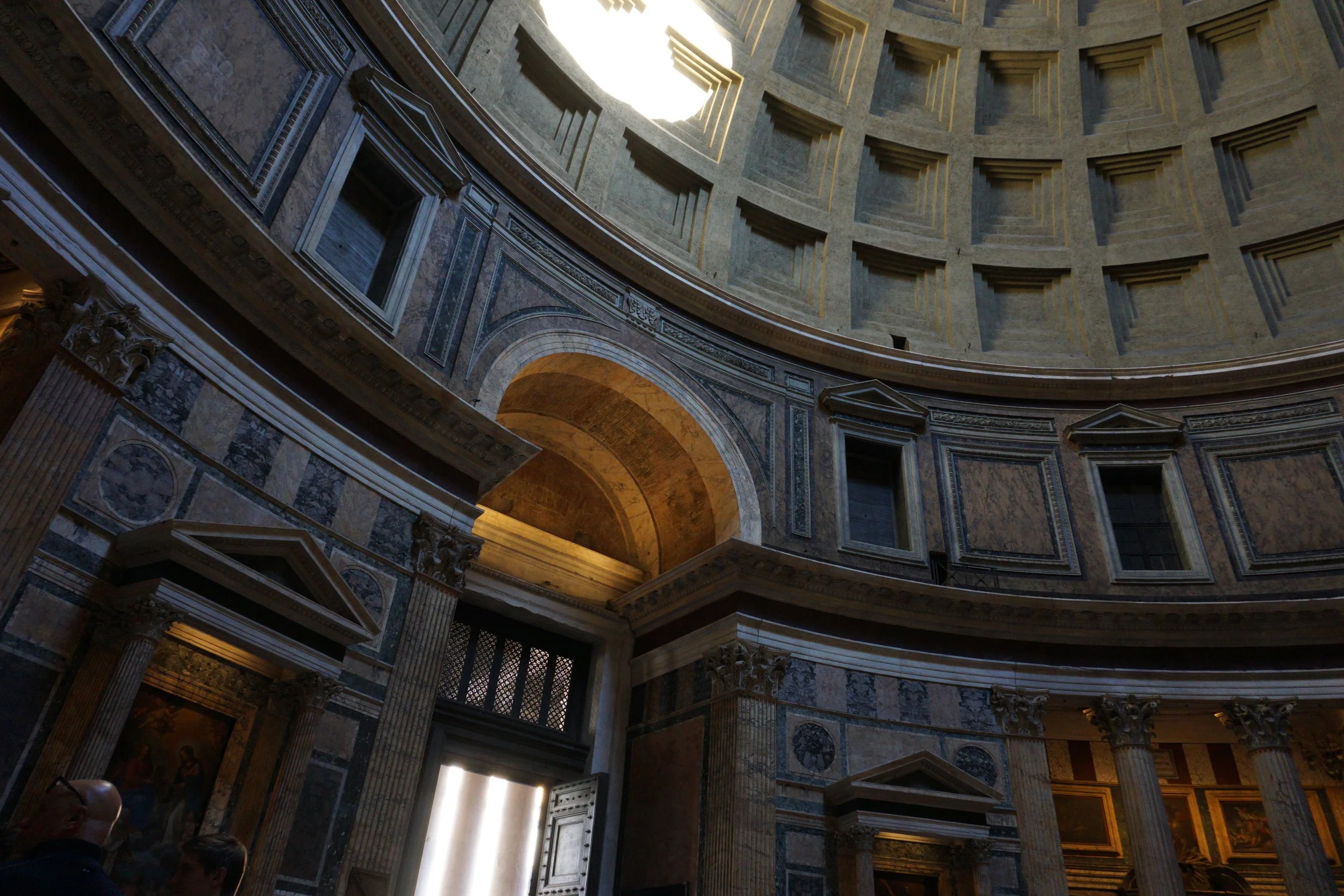Interior view of the Pantheon, showing the oculus at the top of the dome letting sunlight in, with ornate columns, arches, and marble walls.