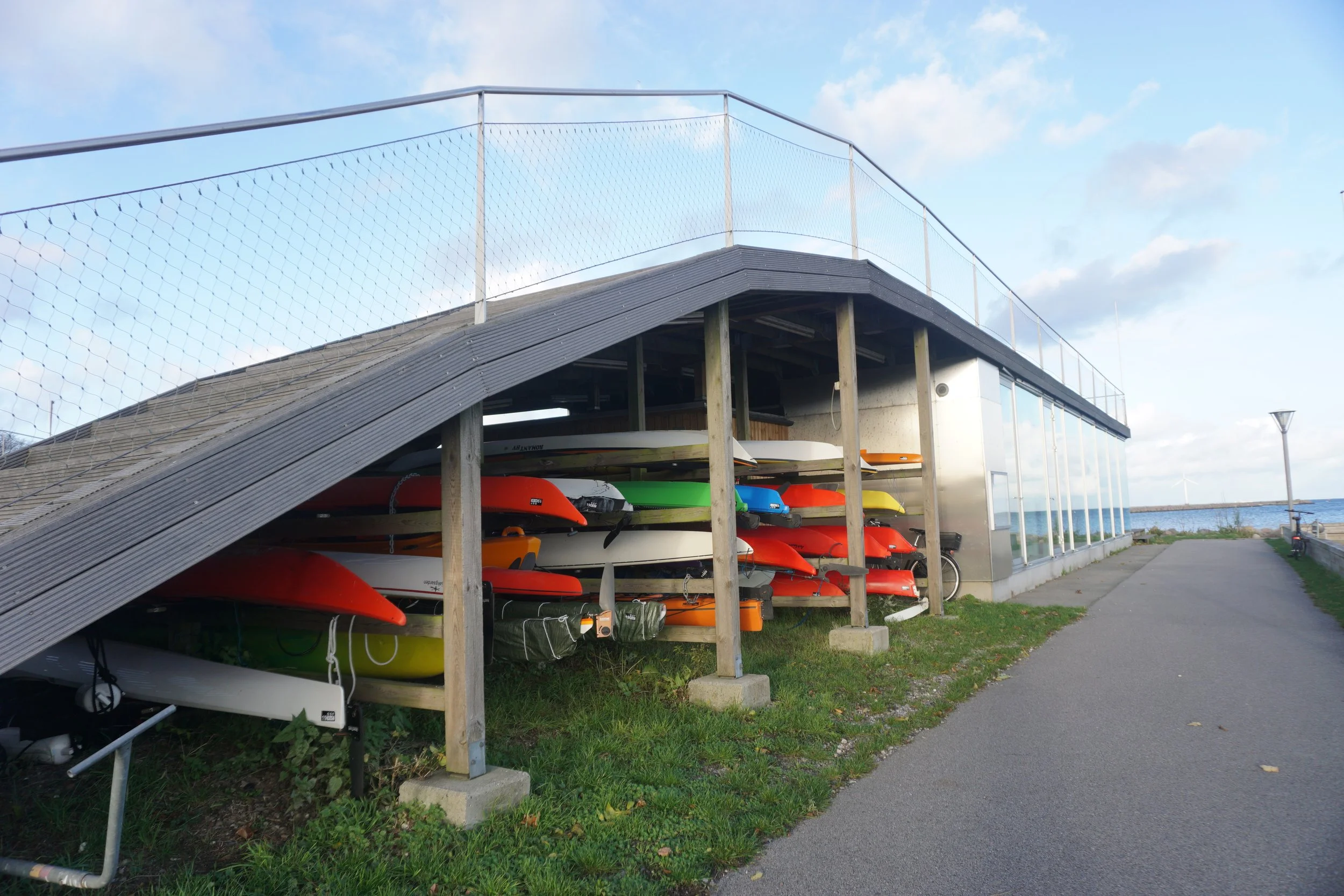 Colorful kayaks stored on racks under a building near a sidewalk, with a body of water and cloudy sky in the background.