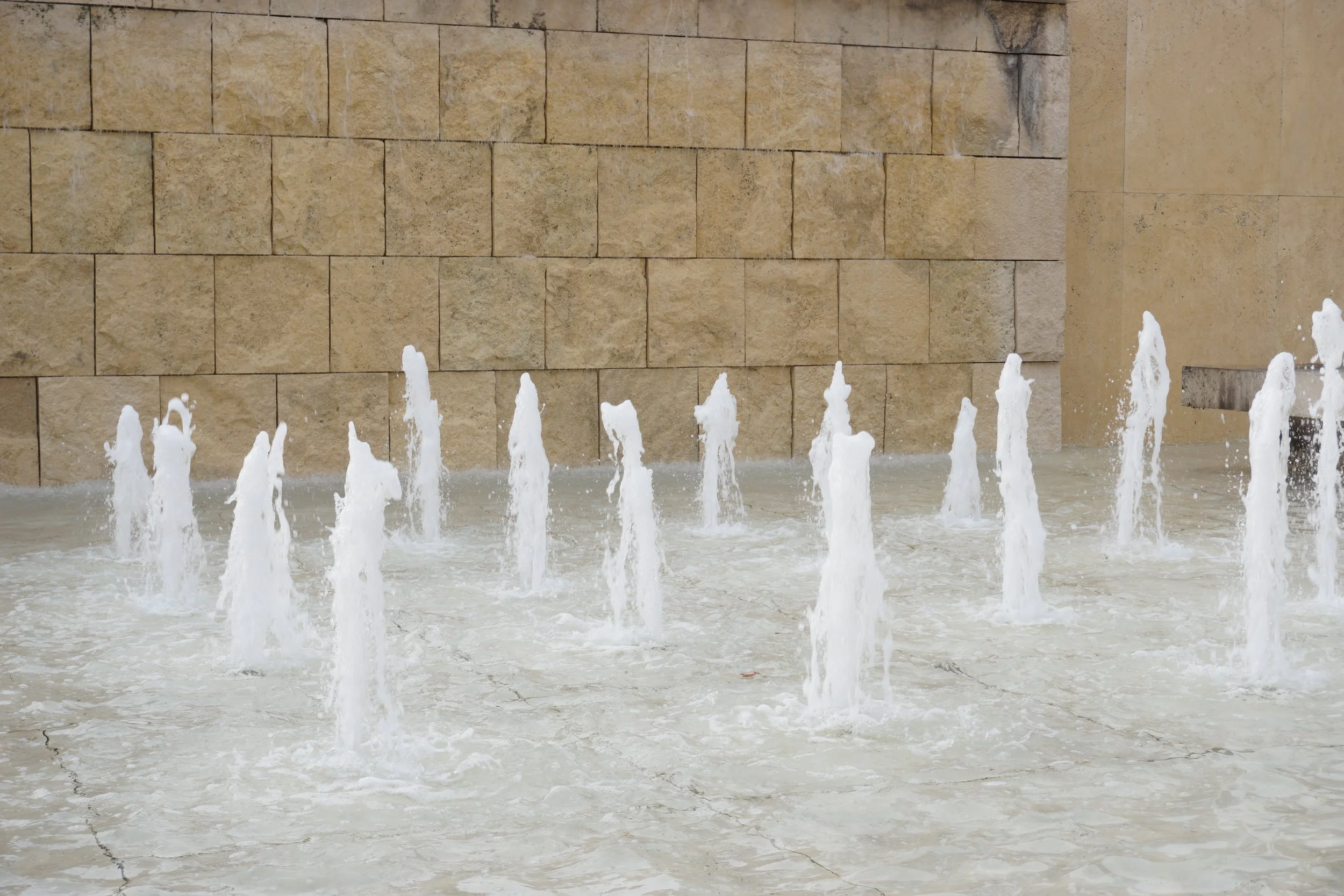 A fountain with multiple vertical streams of water shooting upwards in front of a beige brick wall.