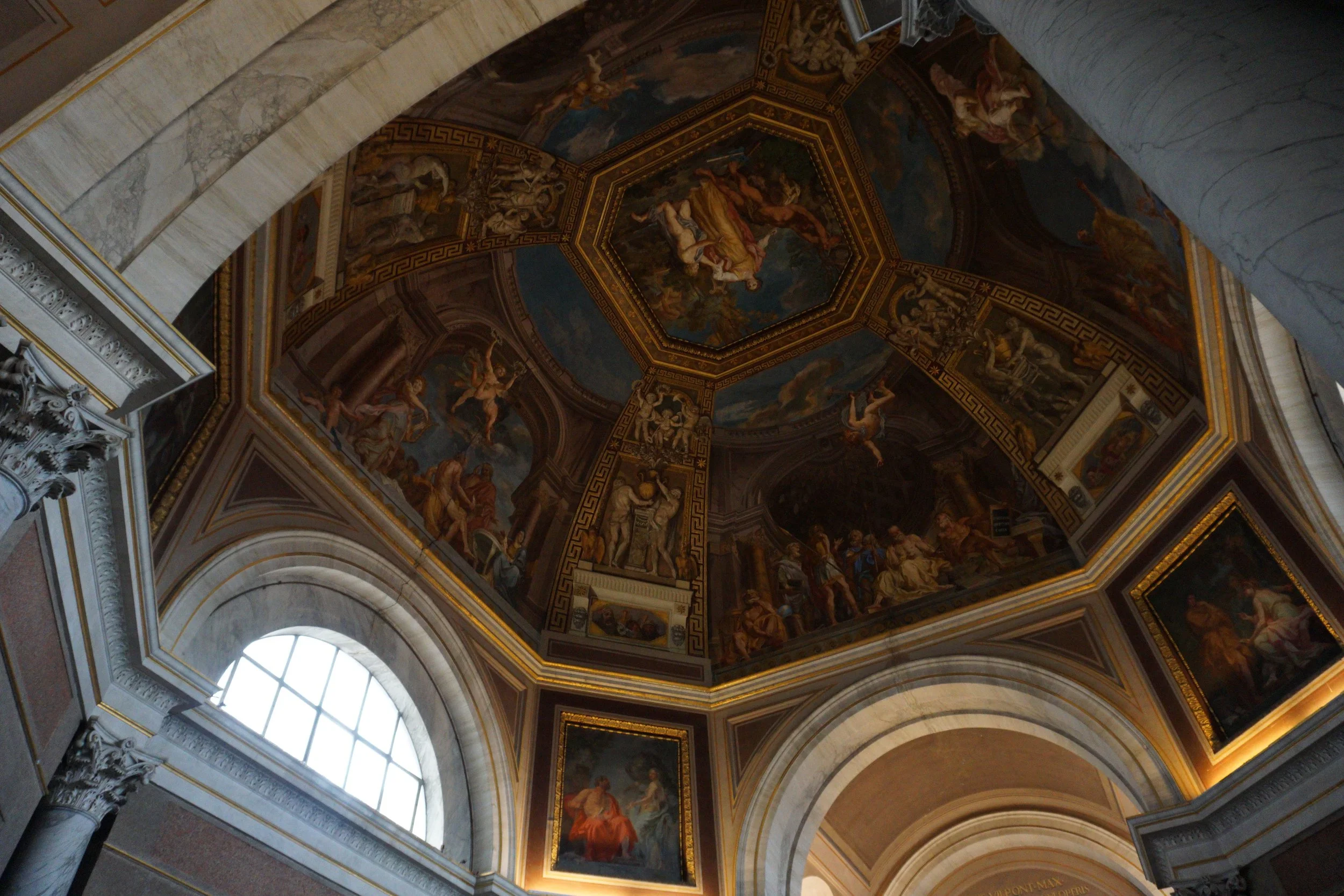 The image shows an ornate ceiling and upper walls of a historical building, decorated with elaborate paintings, sculptures, and gold accents, illuminated by natural light from large arched windows.